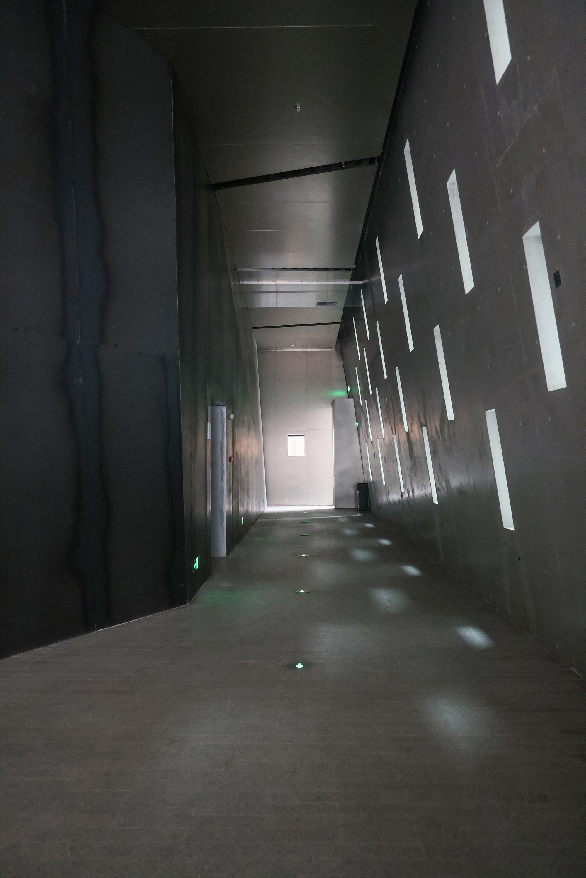 Dark corridor with vertical light slots piercing the concrete wall casting geometric shadows on the floor