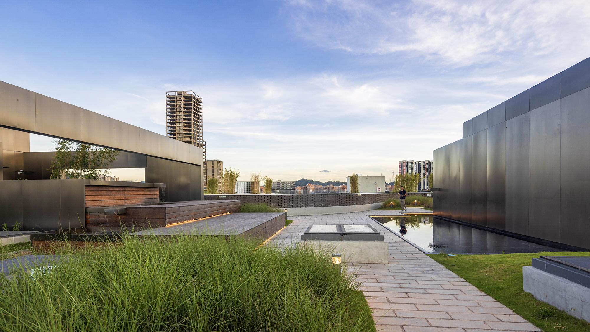 Rooftop terrace with reflecting pool, paved walkways, planted grasses and dark metal perimeter walls at dusk