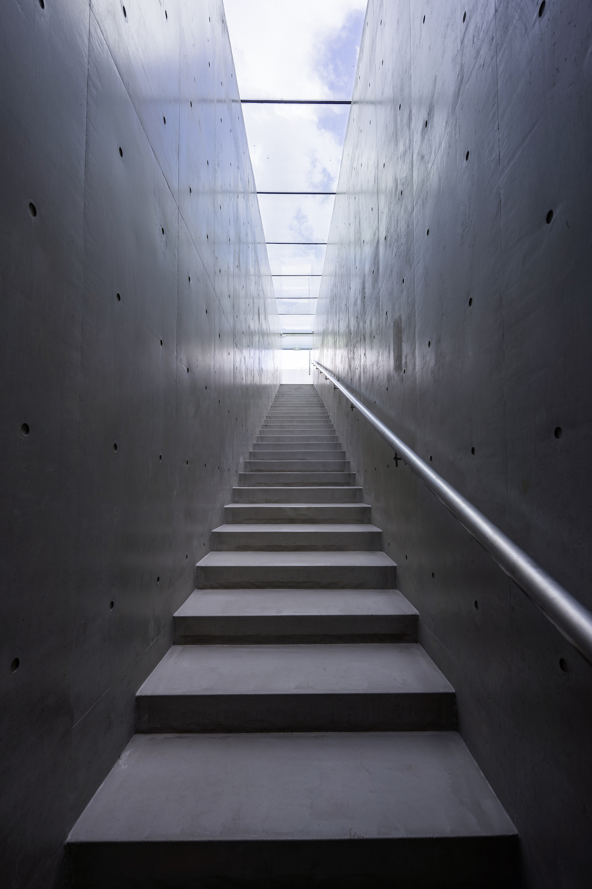 Concrete staircase ascending between tall cast-in-place walls beneath a linear skylight admitting daylight