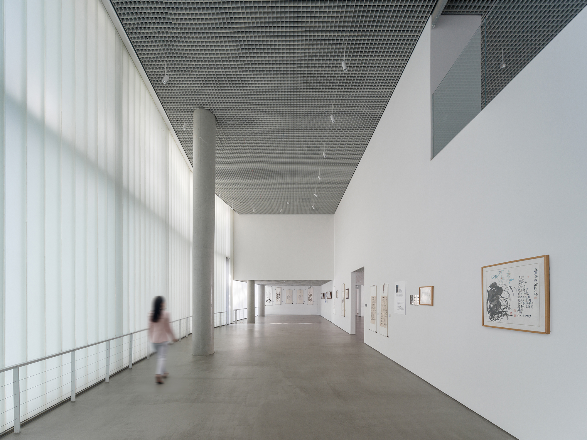Gallery with perforated metal ceiling, translucent curtain wall and a figure walking past framed artworks