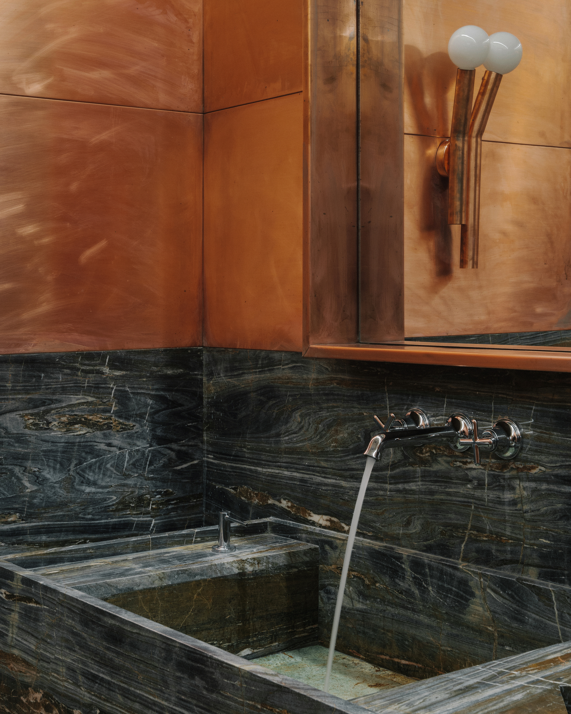 Close-up of dark marble sink with wall-mounted faucet beneath copper panel and sconce