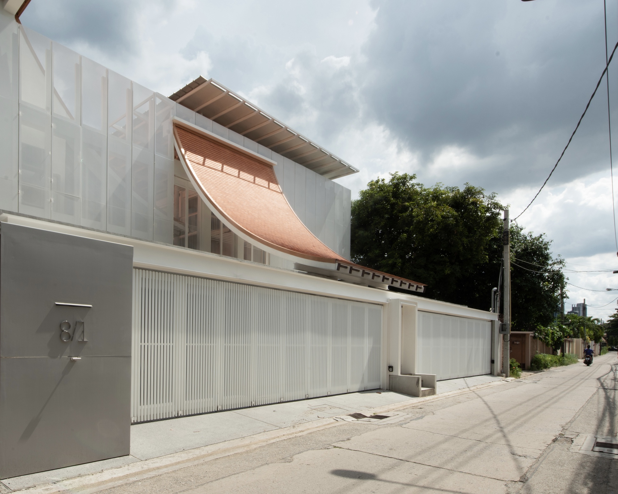 Street view of the white paneled facade with curved clay tile roof element under cloudy skies