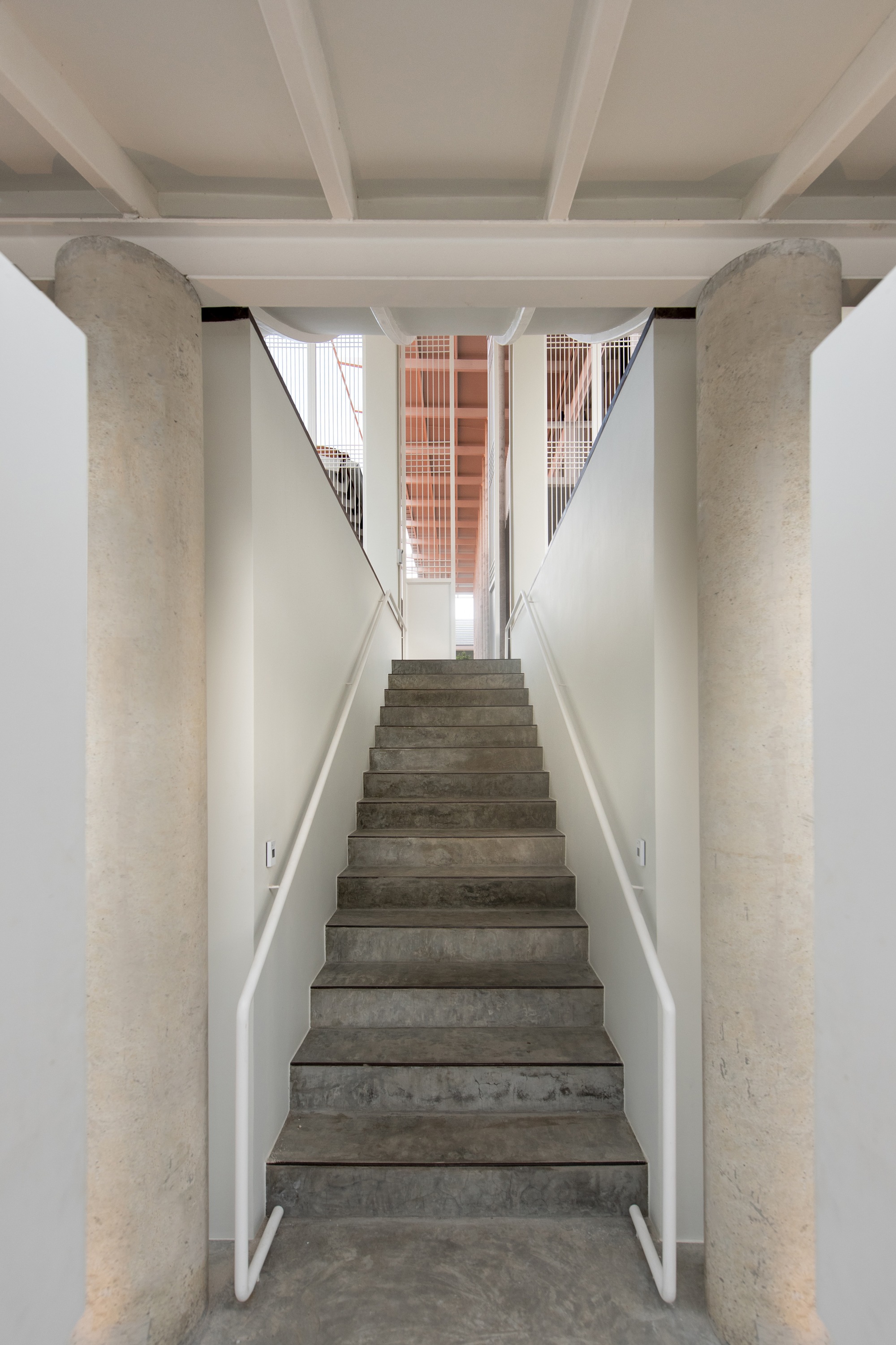 Concrete staircase flanked by cylindrical columns with exposed beams overhead and terracotta screen visible at landing