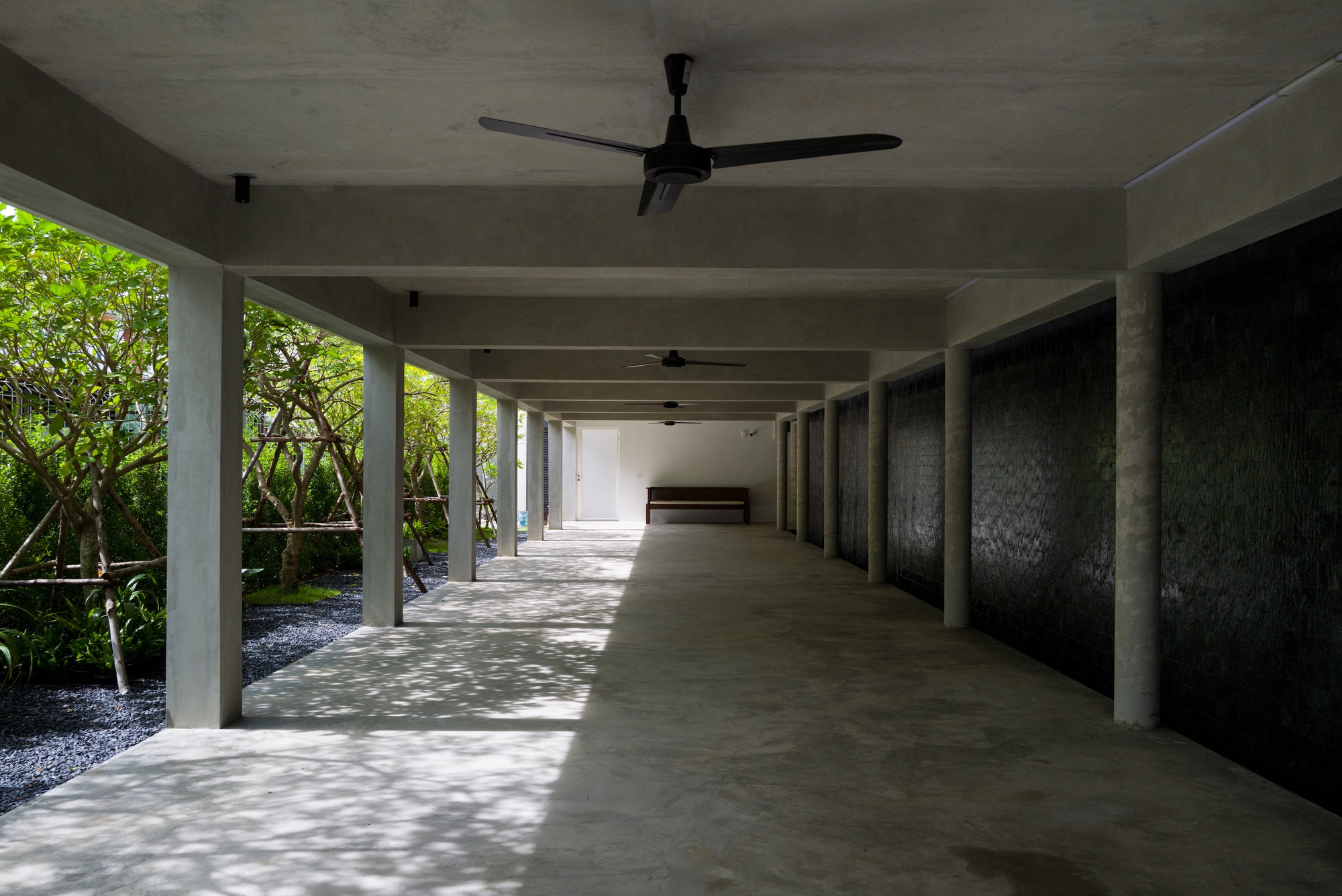Covered corridor with concrete columns and ceiling fans beside planted bed with lush green foliage