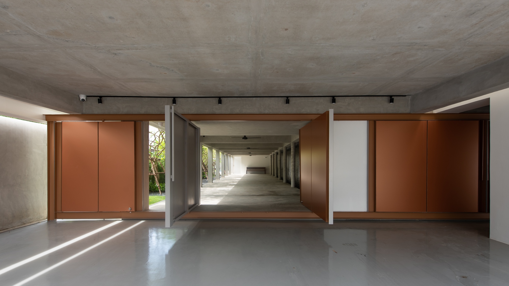 Interior view through large pivoting copper-clad doors opening to a concrete colonnade with sunlight streaming across the polished floor