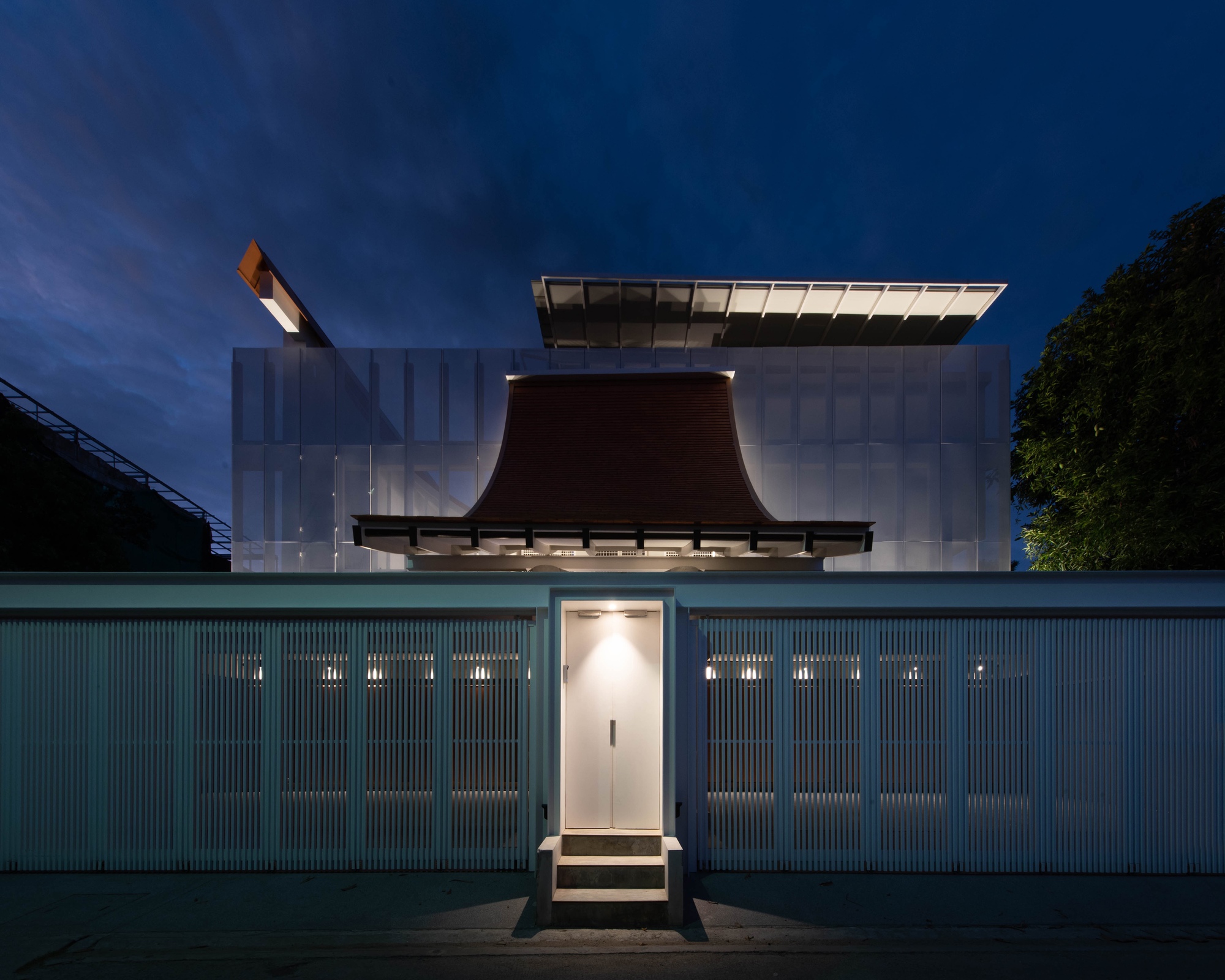 Street facade with corrugated metal fence and curved copper roof element rising above translucent panels at dusk