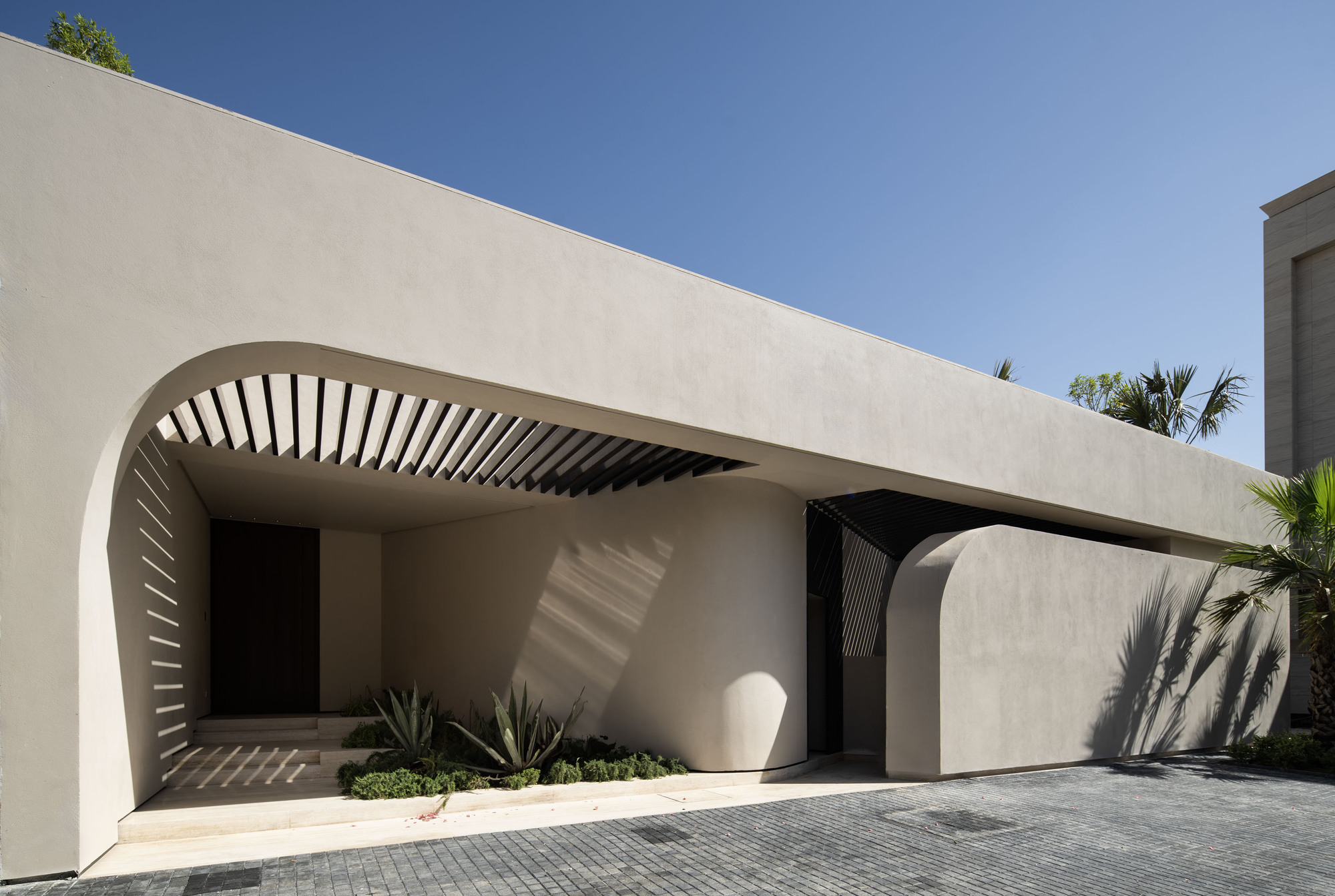 Arched entry with radial timber slat canopy casting shadows on pale stucco wall and planted bed