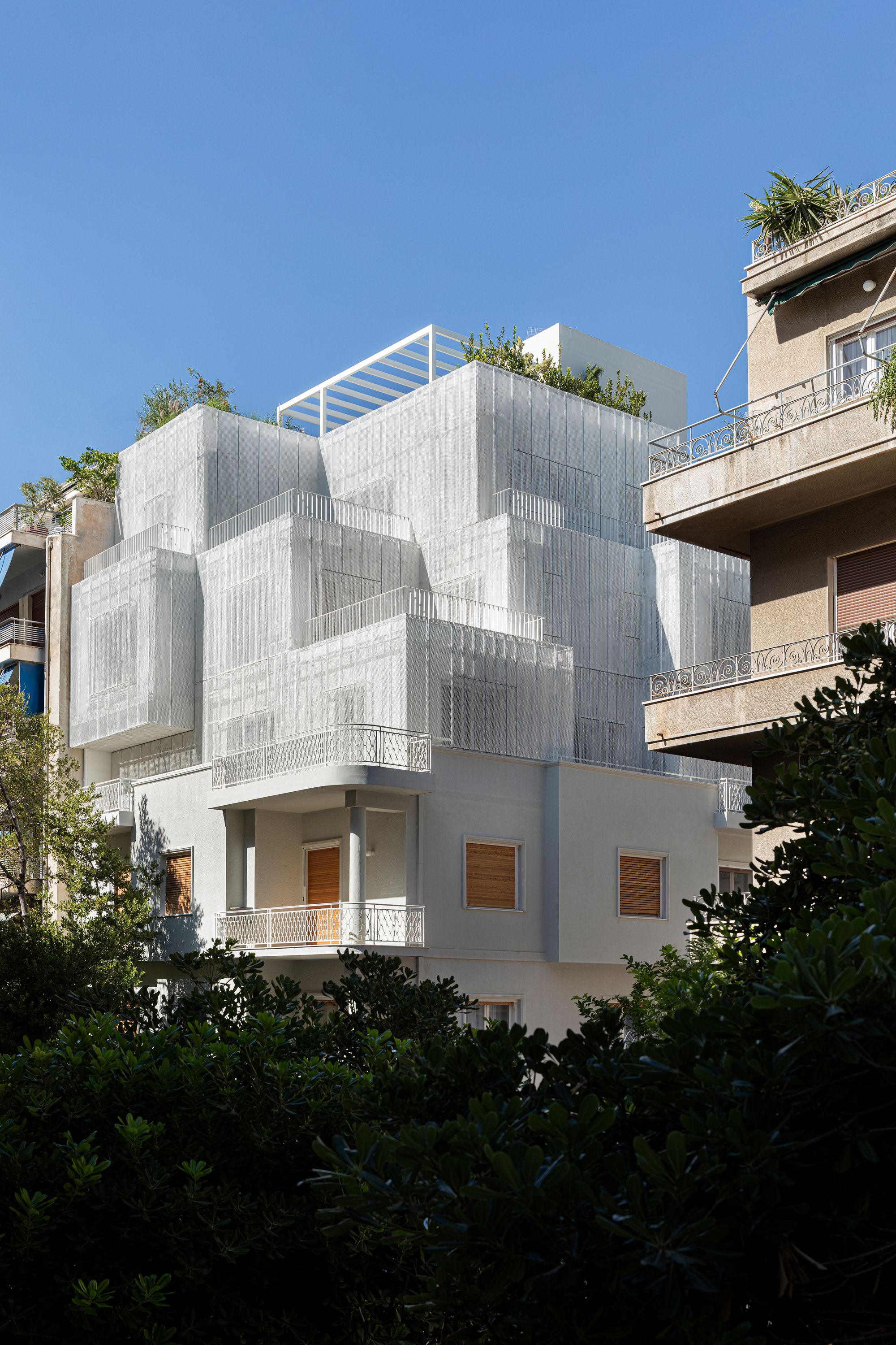 Street view of the layered white metal screen facade with rooftop garden framed by neighboring buildings