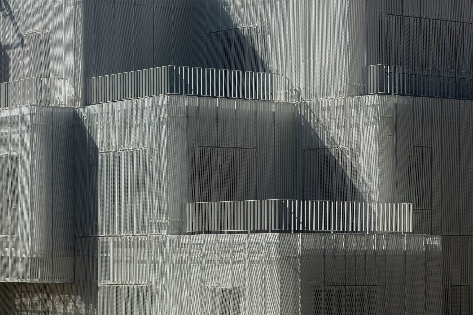 Offset balconies with slatted railings recessed into perforated metal screen facade with shadow patterns across surface