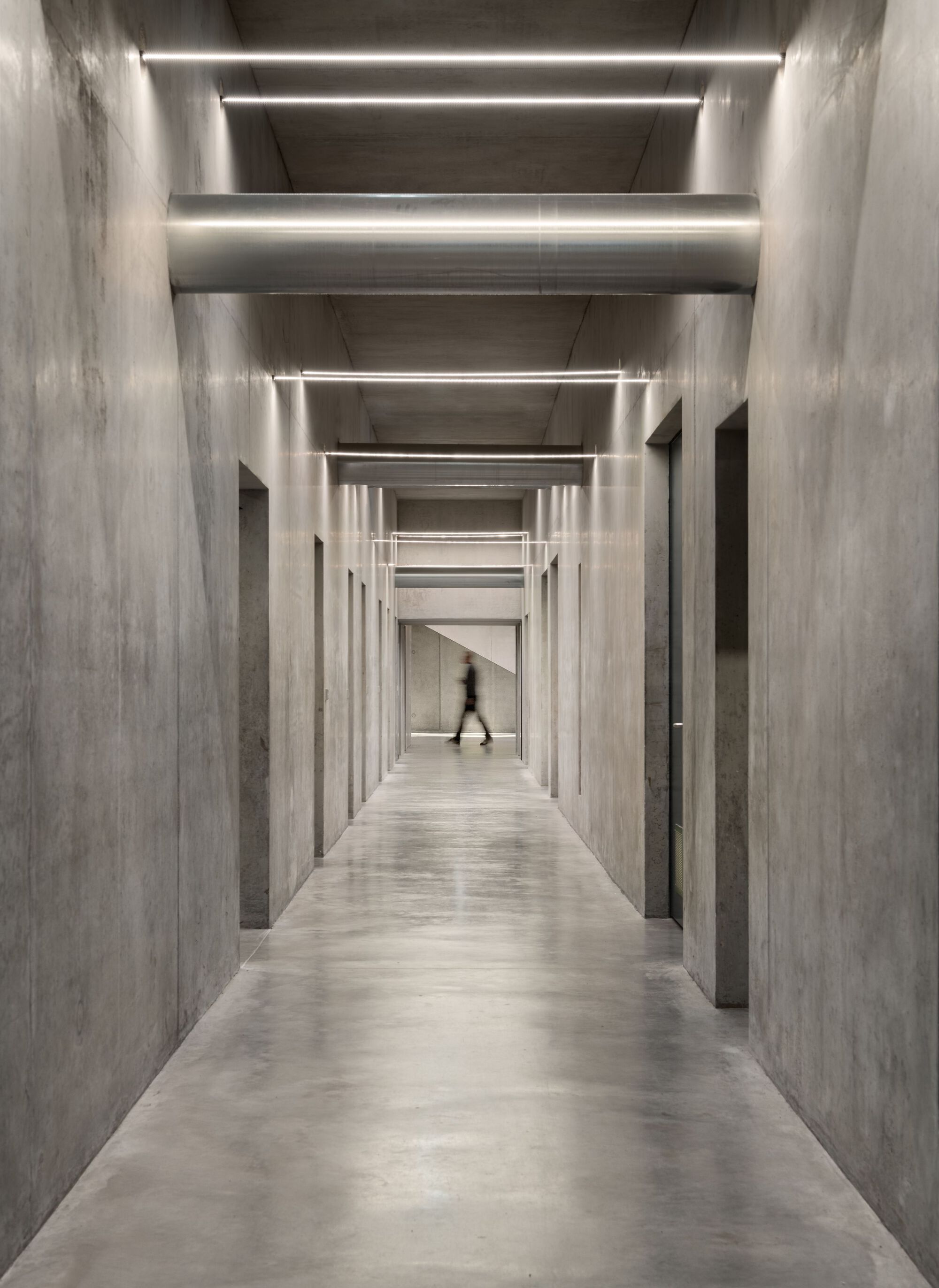 Concrete corridor with linear ceiling lighting and a figure walking through recessed doorways