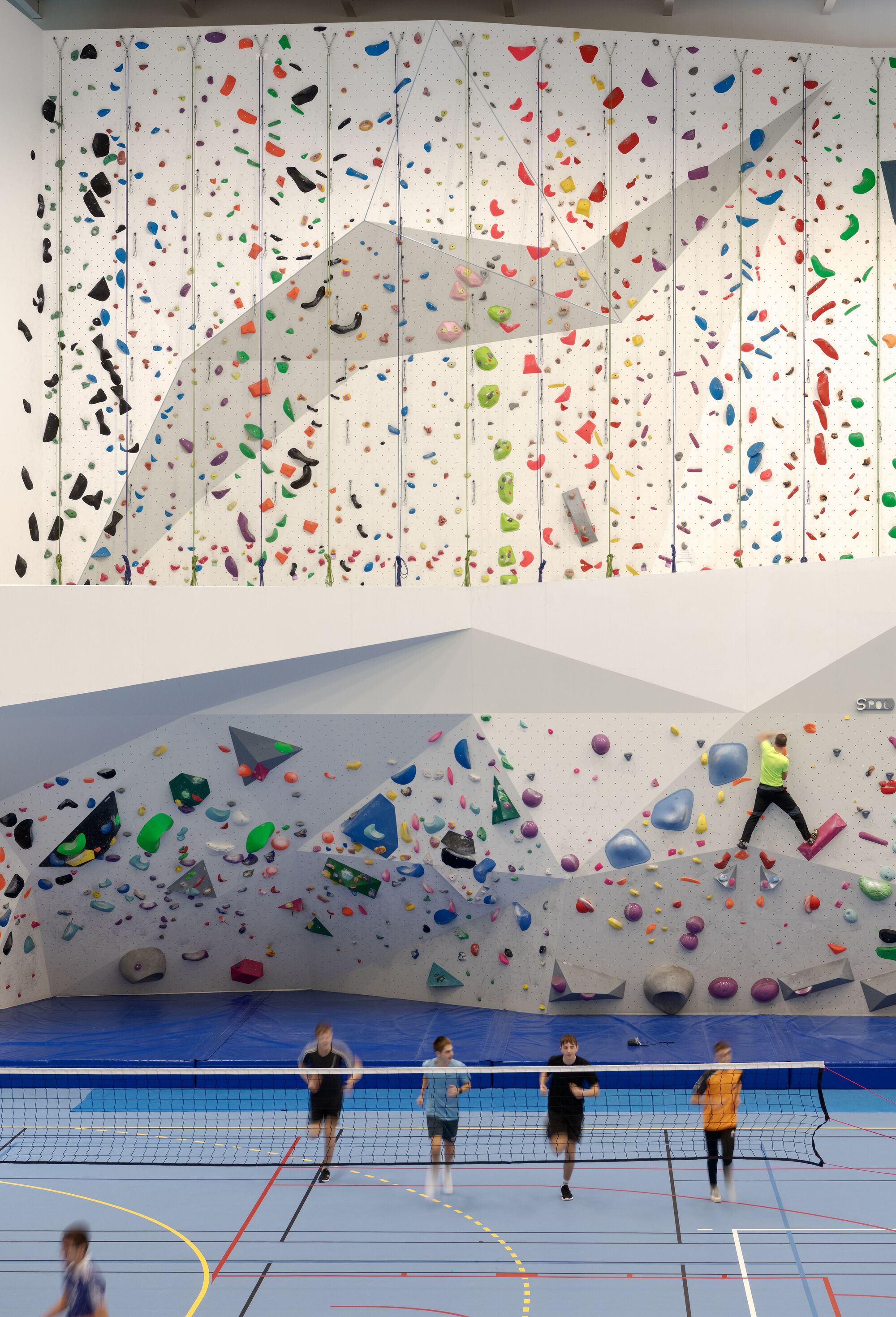 Four people standing at badminton net below two-level climbing walls with multicolored holds