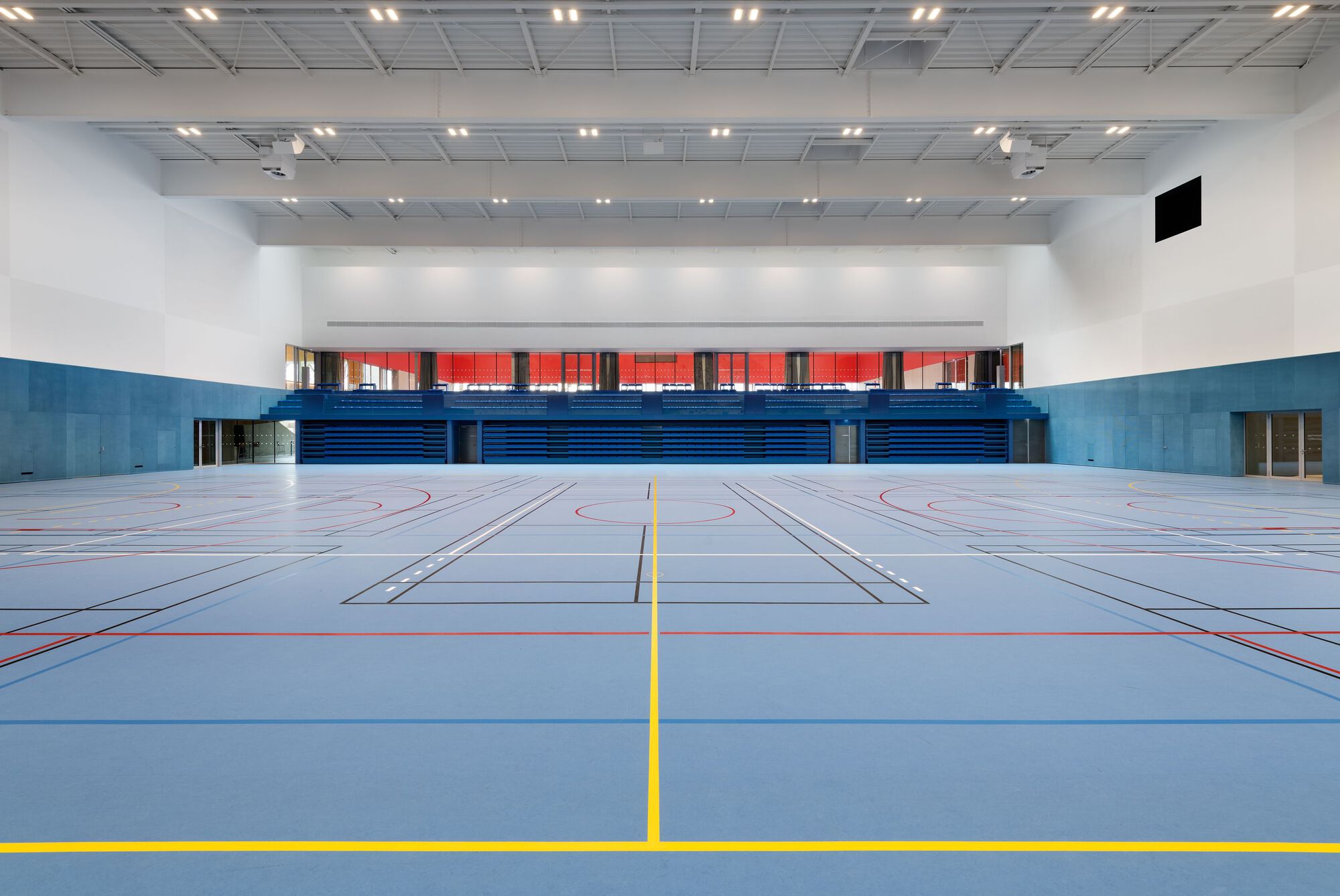Interior multipurpose sports hall with blue tiered seating and a marked floor under recessed ceiling panels