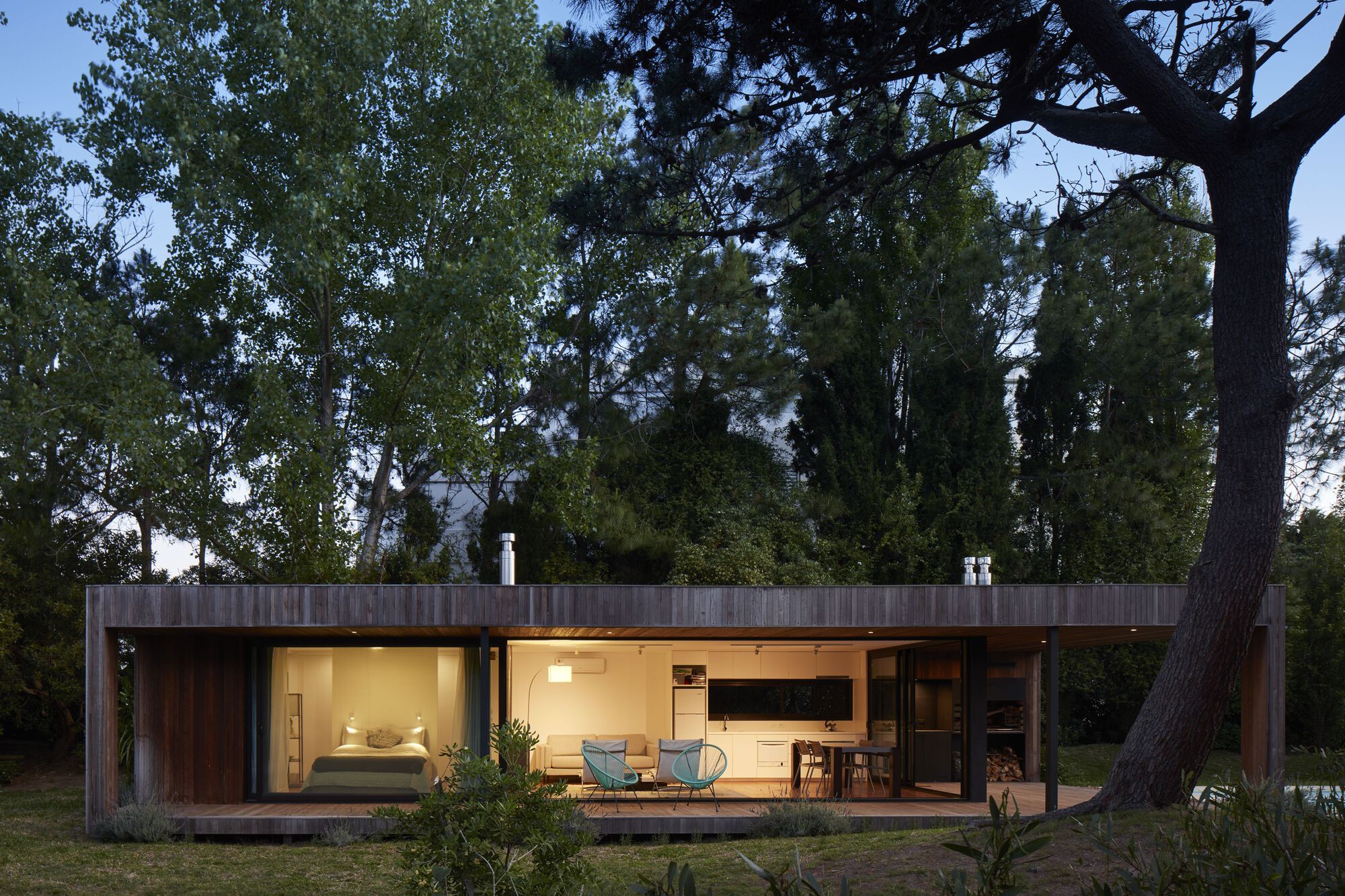 Long timber volume with continuous glazing and lit interior at dusk in a wooded clearing