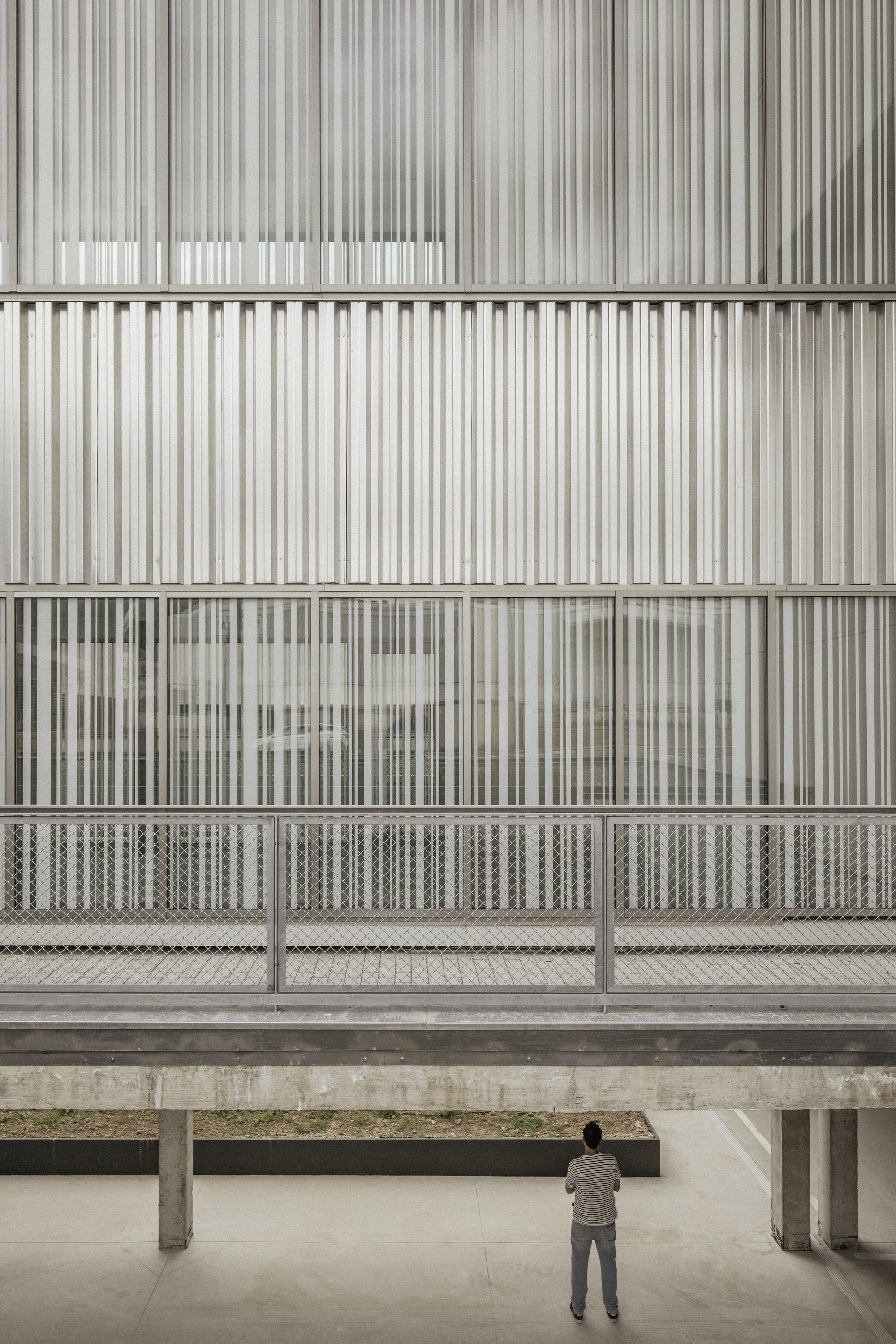Vertical metal screen facade with mesh balustrade viewed from concrete-columned ground-level courtyard with visitor