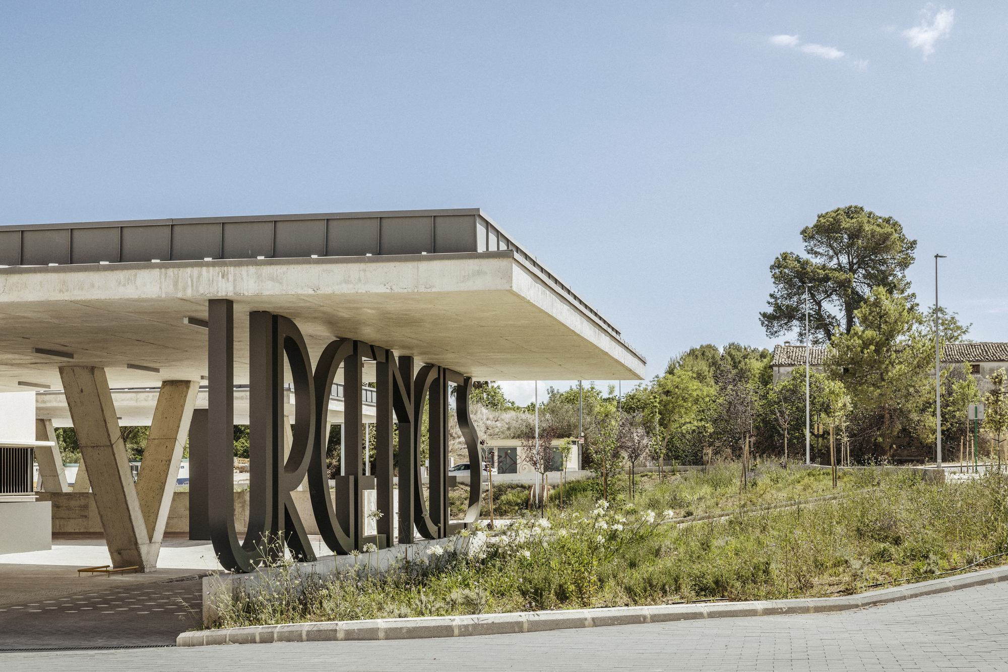 Cantilevered concrete canopy on angled supports with metal lettering and wildflower meadow under blue sky