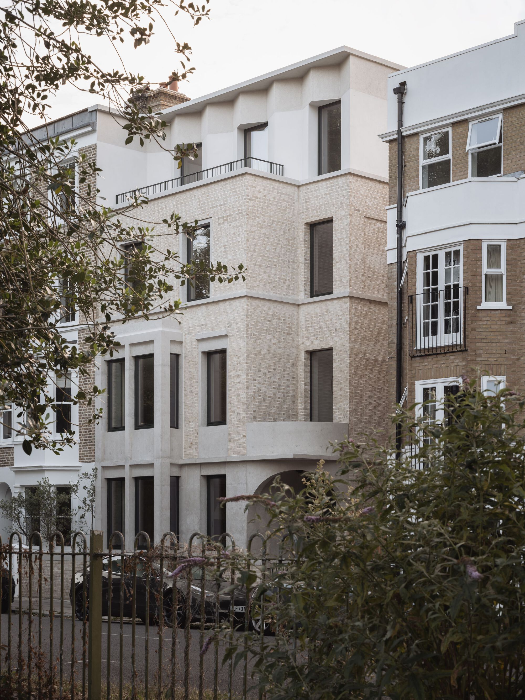 Street view of the pale brick facade with vertical windows nestled between adjacent buildings and greenery