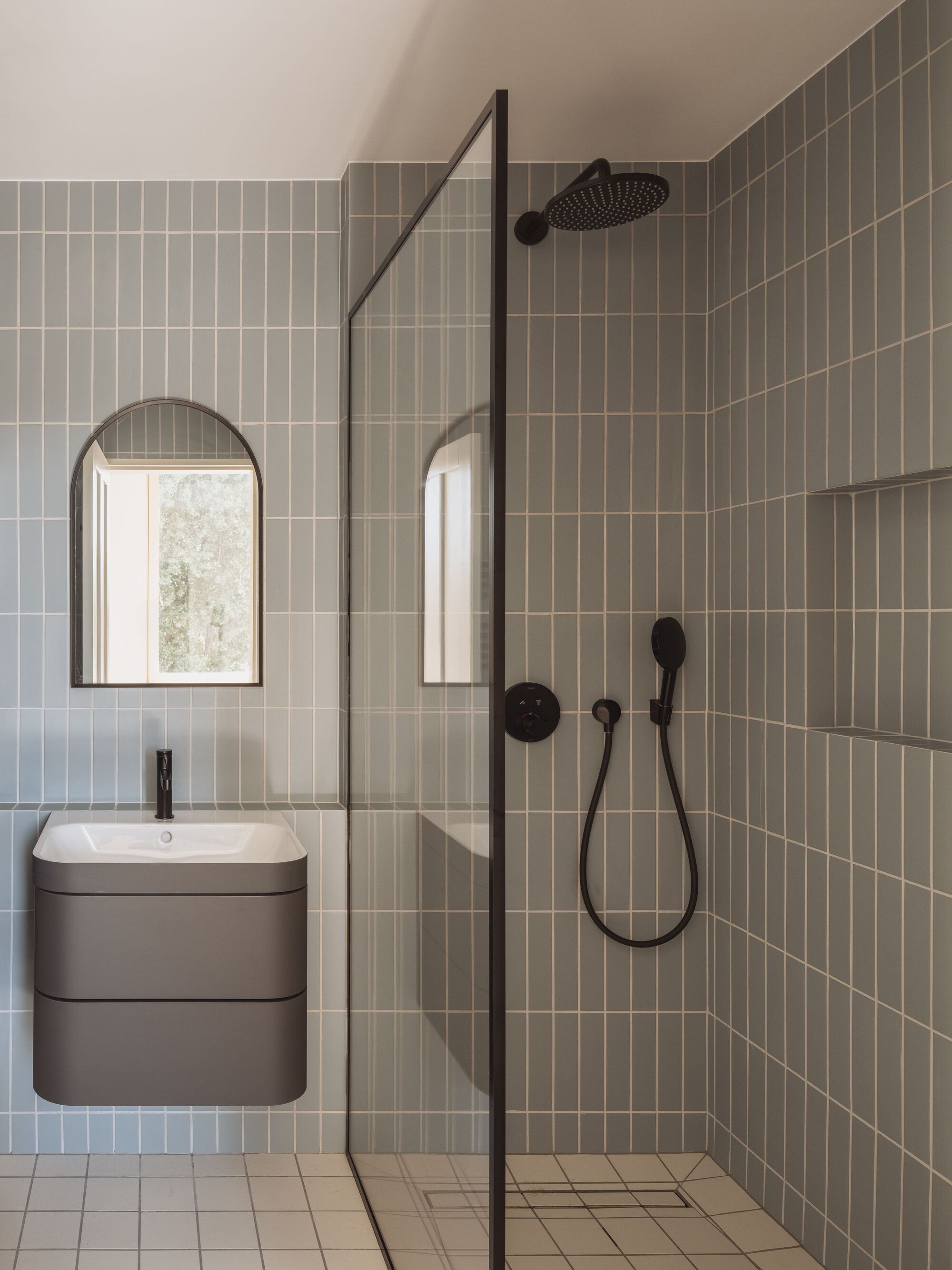 Bathroom interior with olive vertical tile shower enclosure, arched mirror and wall-mounted vanity under soft daylight