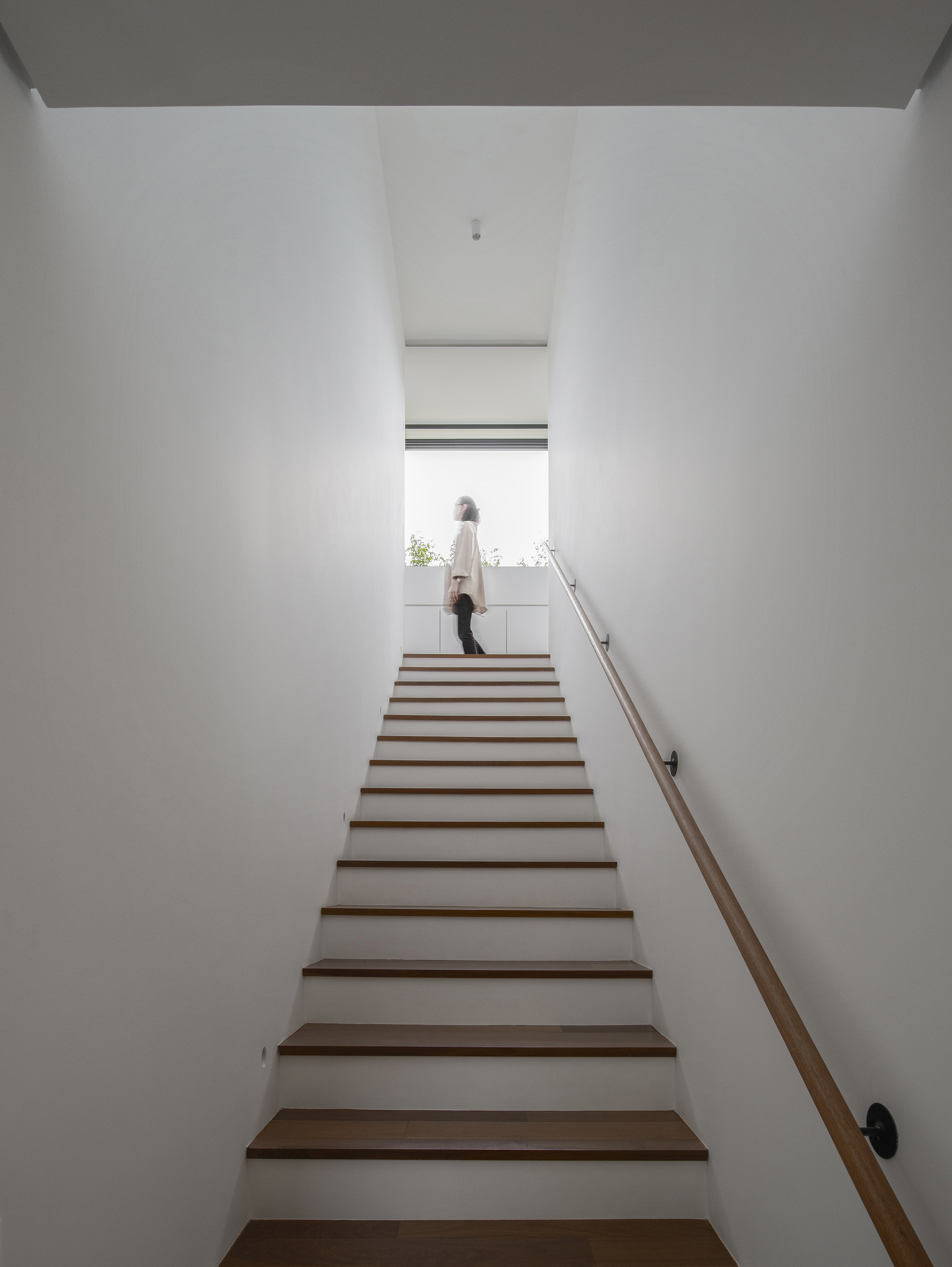 Timber staircase rising between white walls with a figure at the top landing