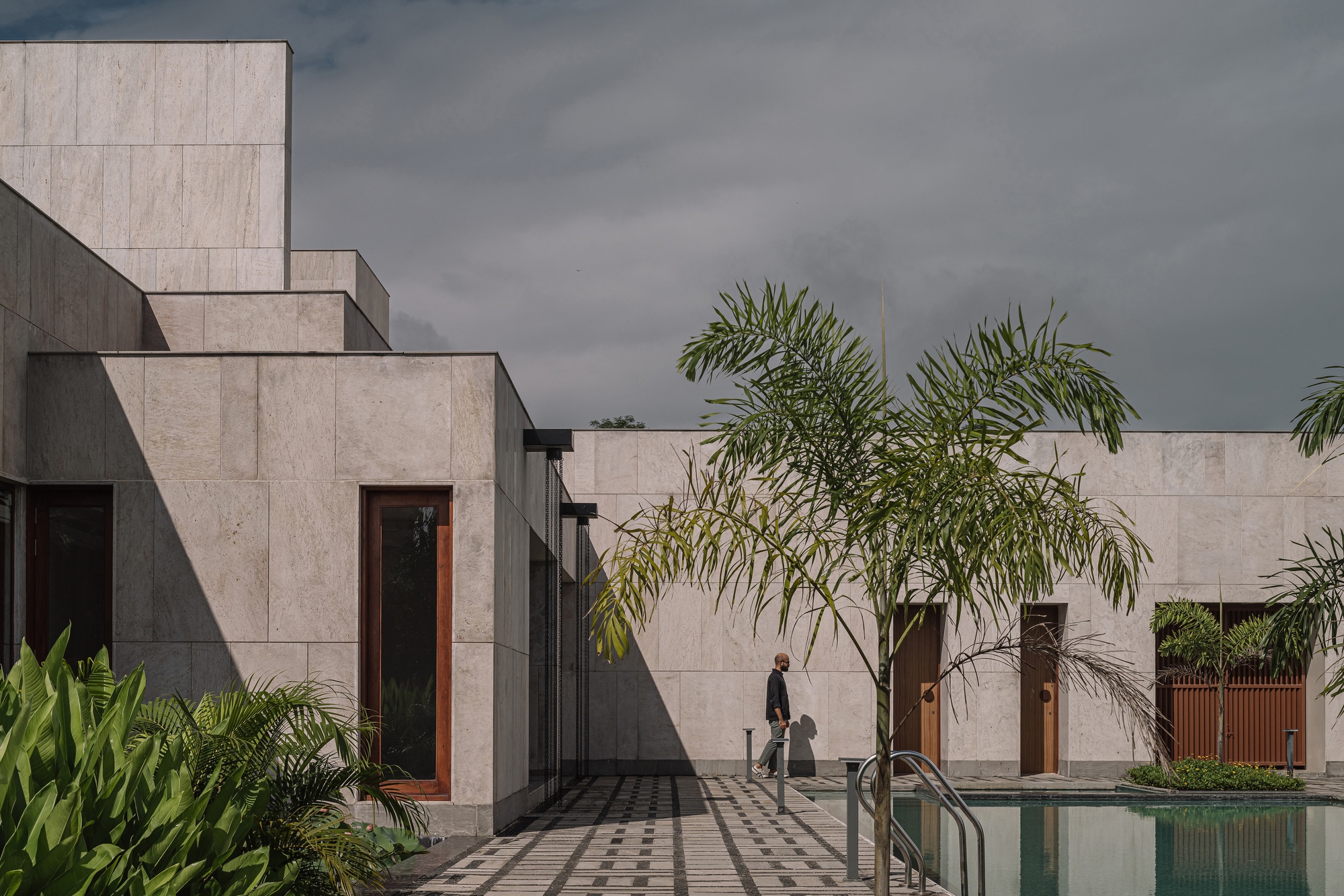 Courtyard view showing concrete volumes, palm trees, and a figure walking near the pool
