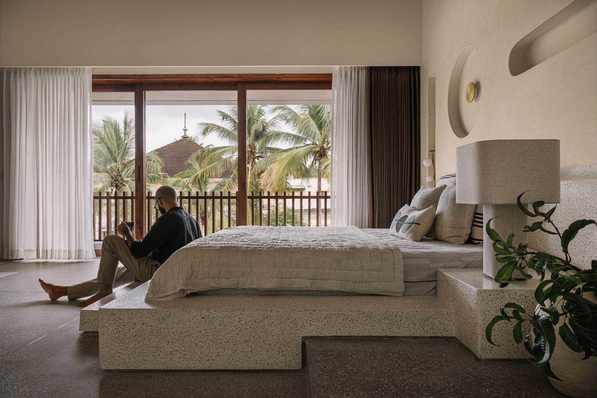 Bedroom with person seated on terrazzo bed platform overlooking palm trees through glazed opening