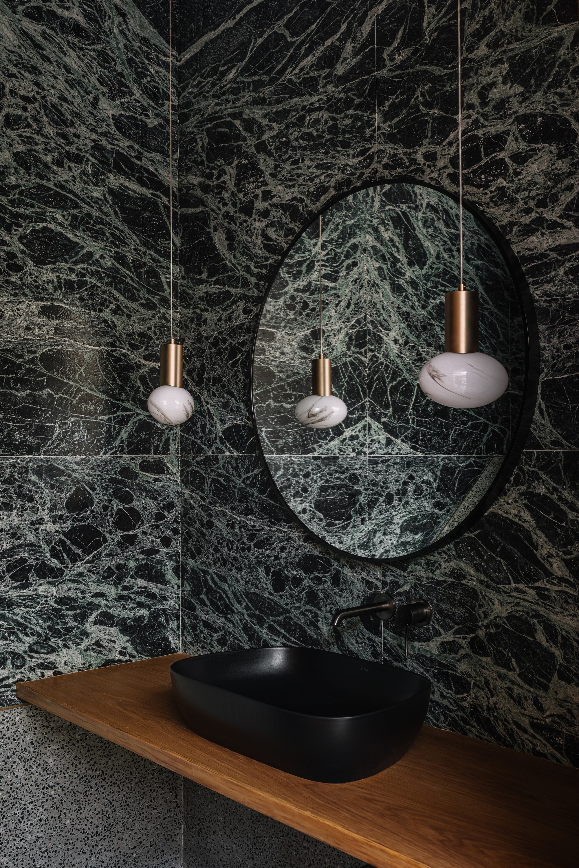 Round mirror reflecting pendant lights above a timber vanity with black basin against green marble