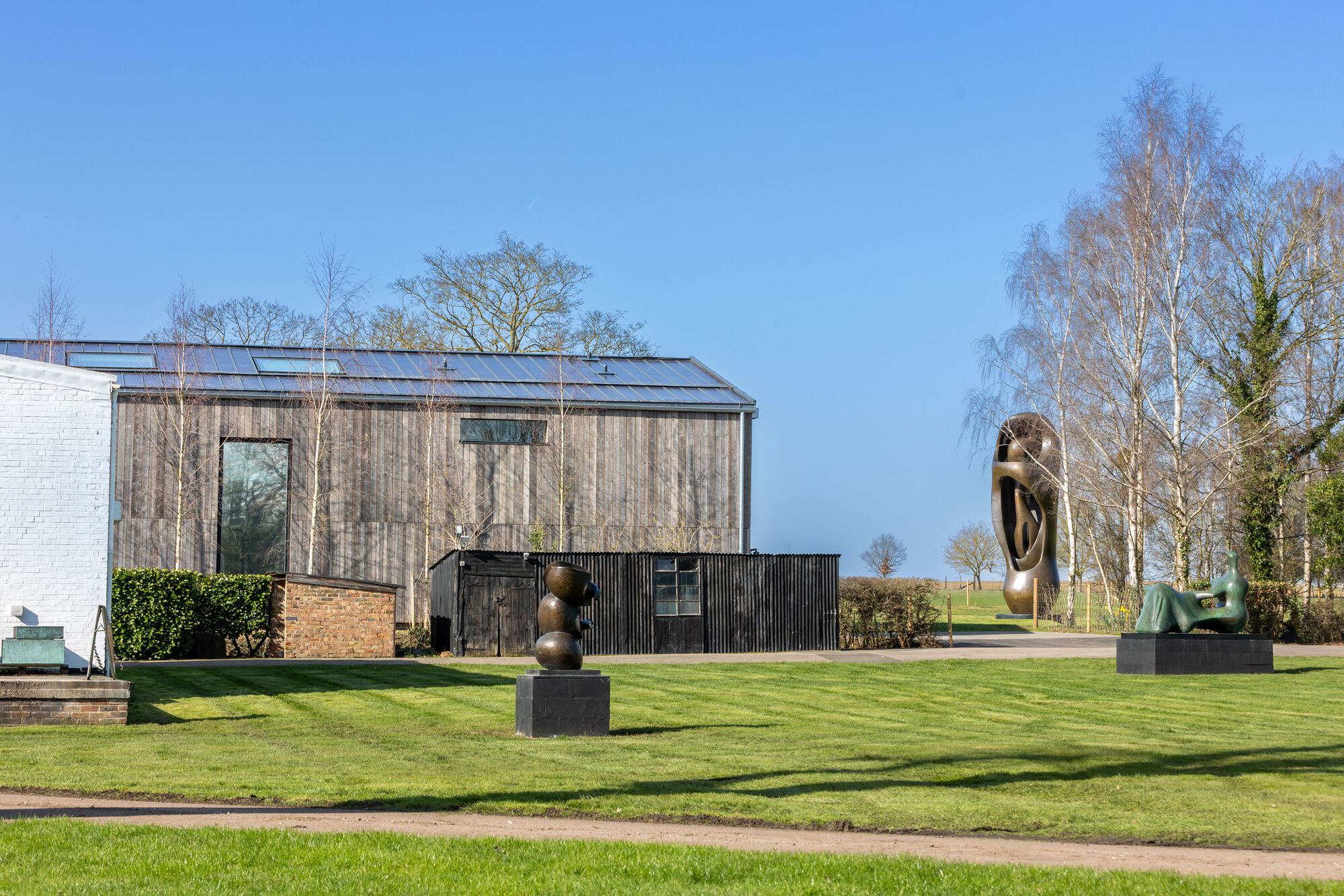 Timber and brick volumes beside a manicured lawn with bronze sculptures and birch trees