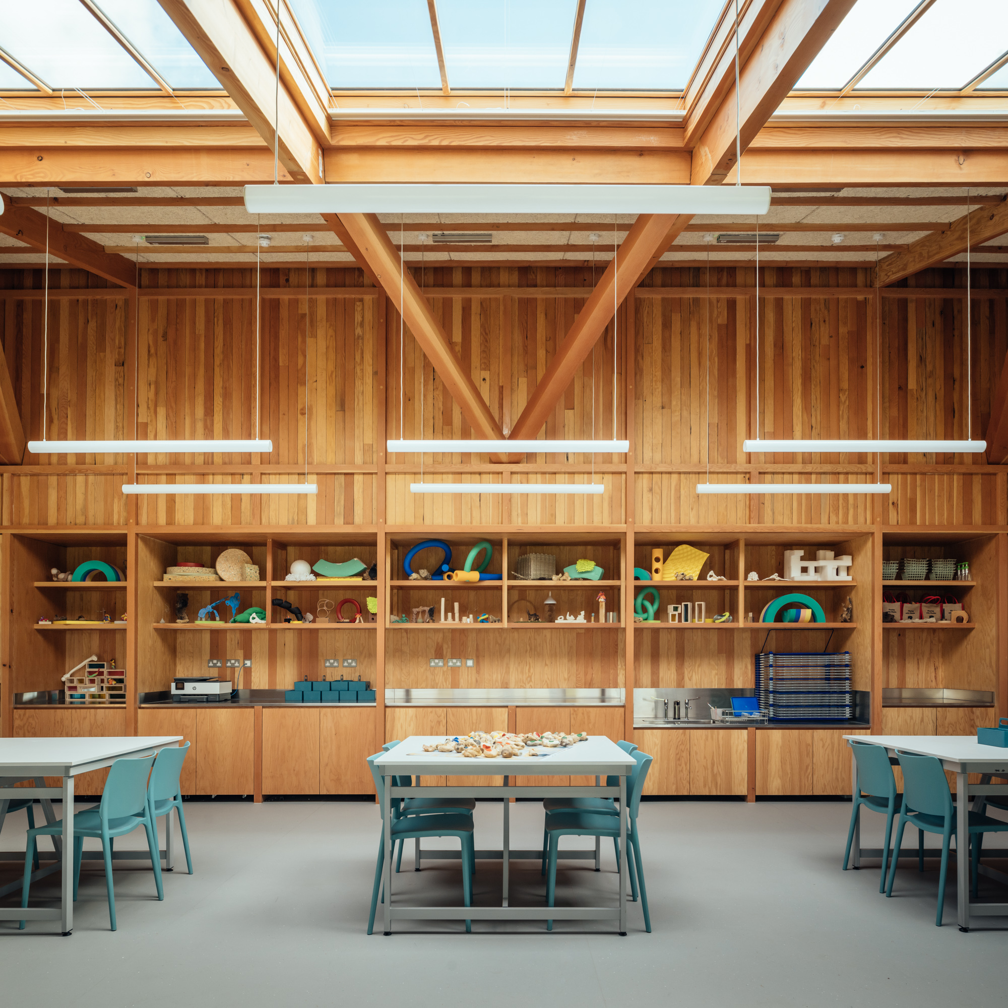 Storage wall with timber shelving beneath exposed beams and continuous roof glazing with suspended light fixtures