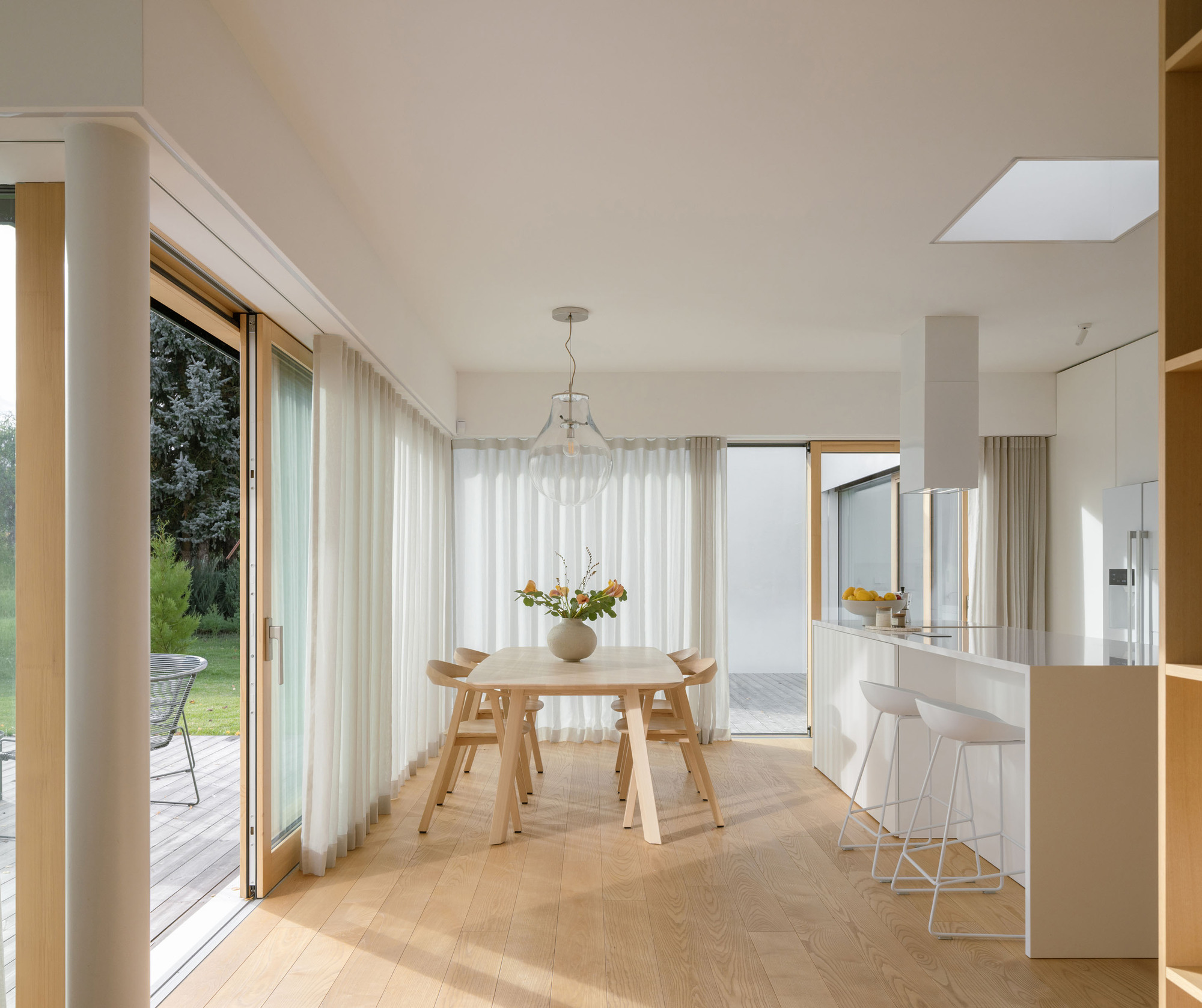 Open dining area with timber table, sheer curtains, and light oak flooring connecting to garden through corner glazing