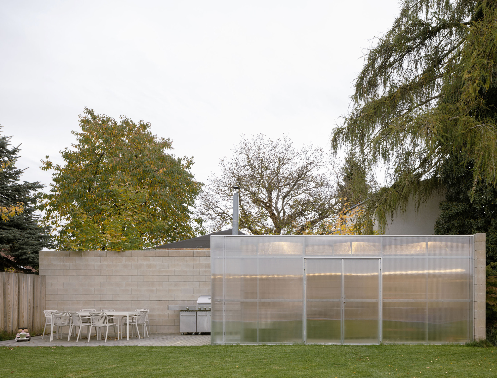 Outdoor dining terrace with concrete paving beside translucent polycarbonate wall and weathered timber fence under overcast sky