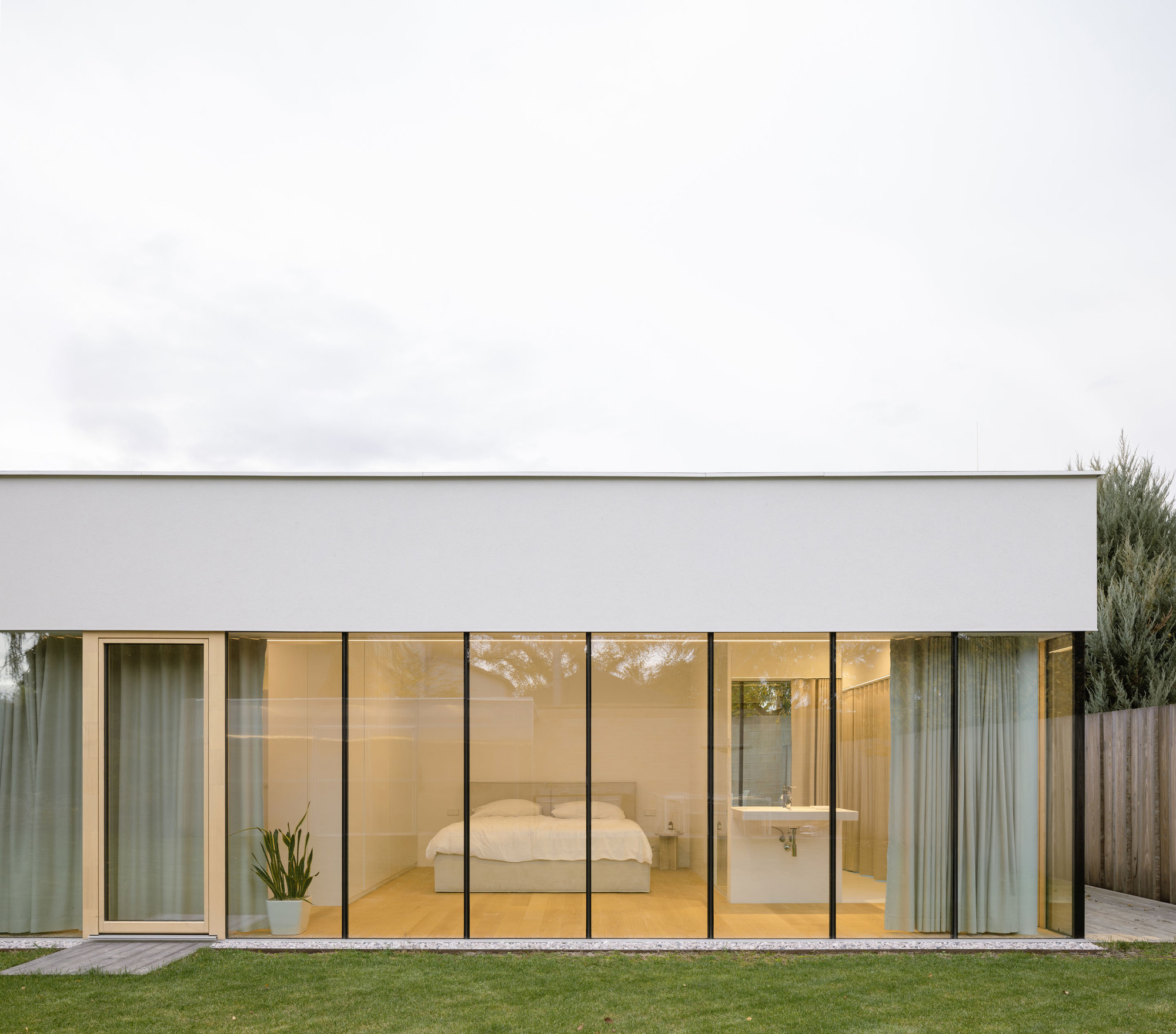 Floor-to-ceiling glazing revealing a bedroom and ensuite bathroom flanked by sheer curtains and lawn