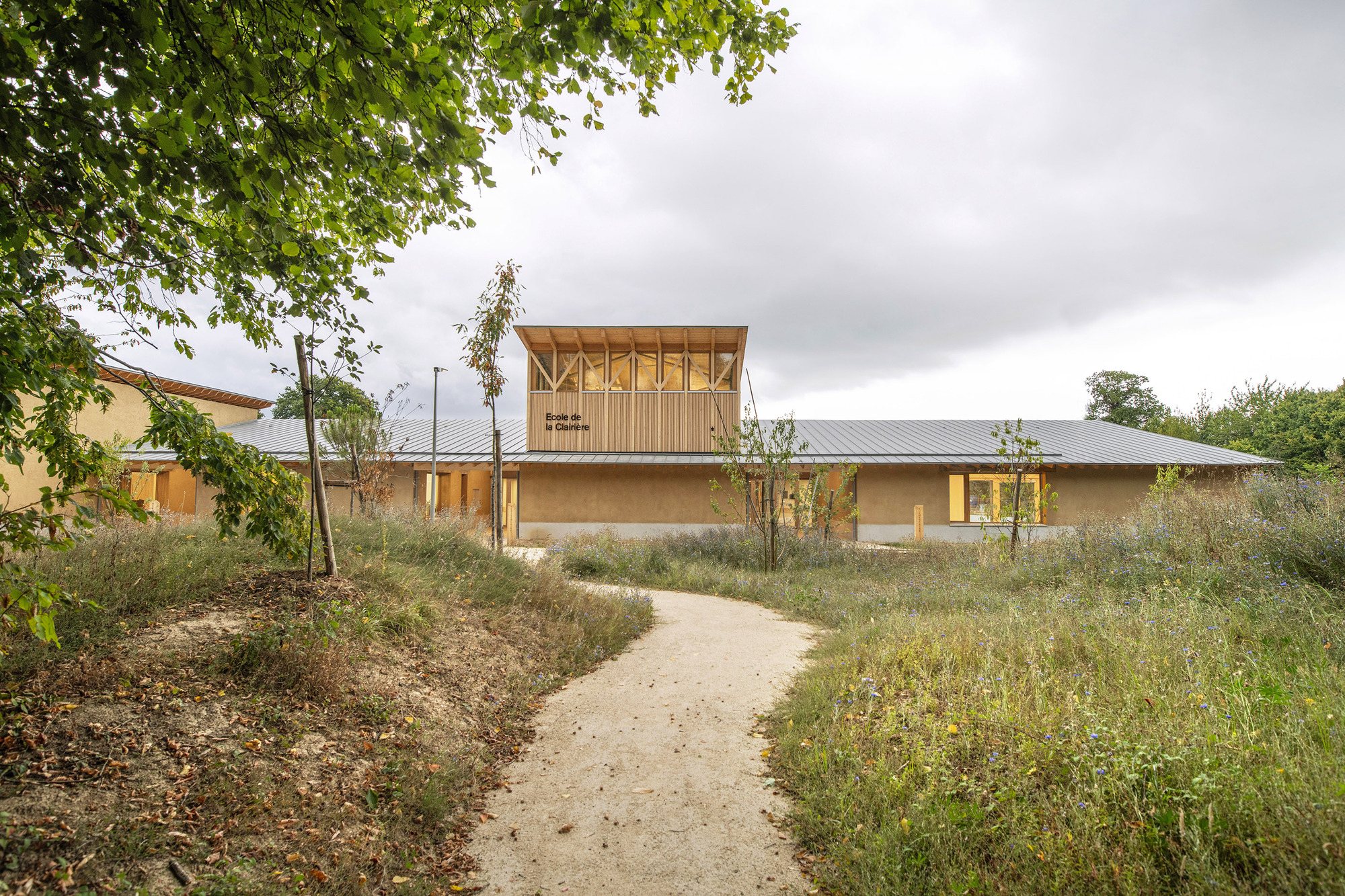 Gravel pathway through wildflower meadow leading to low timber and earthen volumes under overcast sky