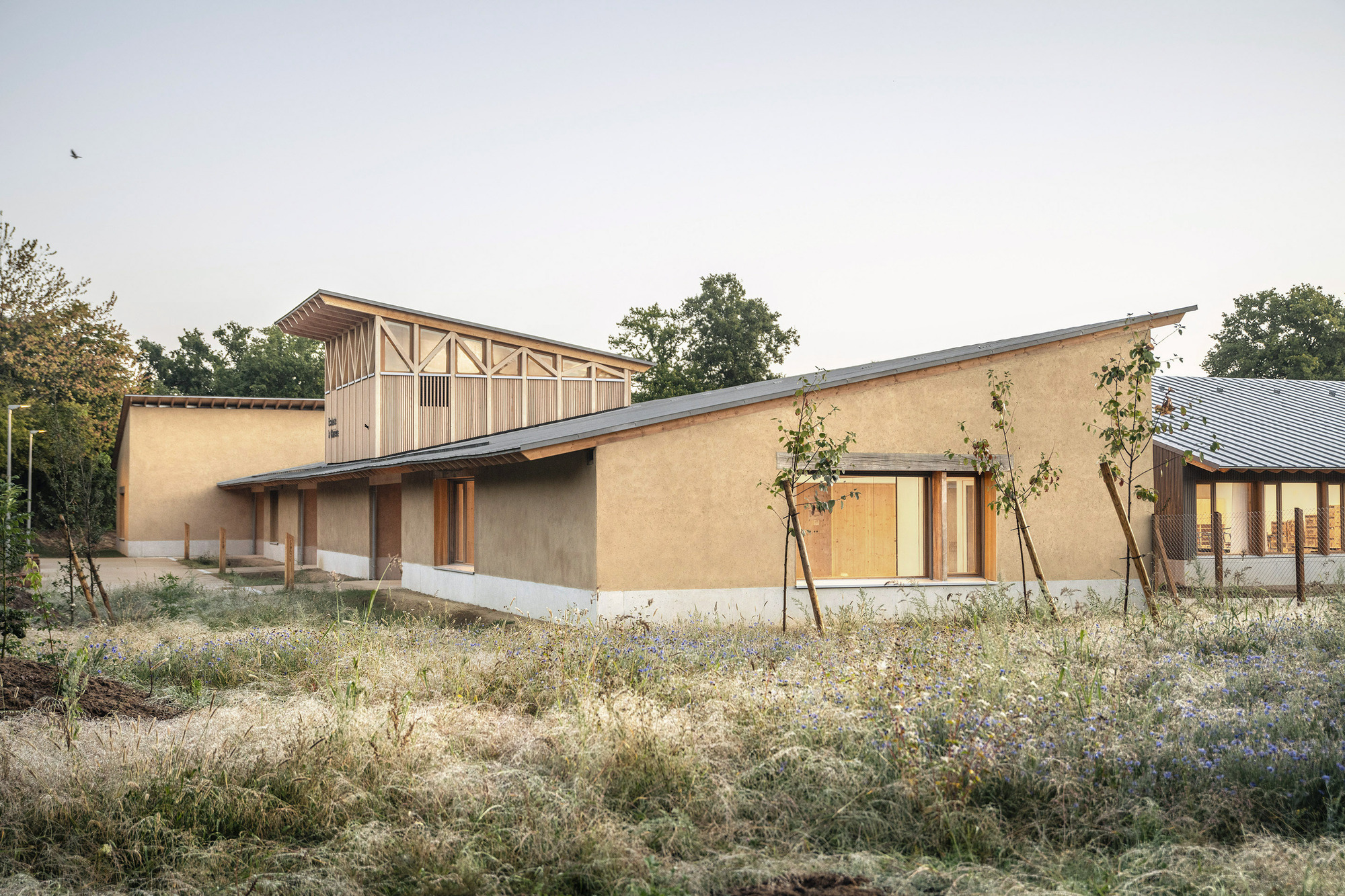 Earthen wall facade with timber columns and glazed openings beside raised timber structure at dusk