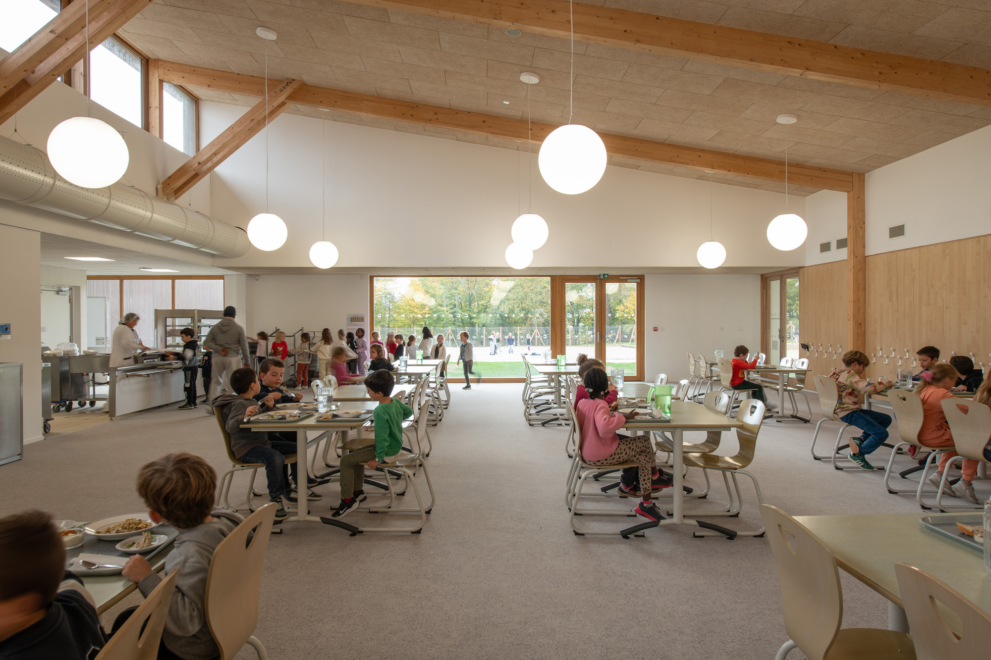 Dining hall with exposed timber rafters, sphere pendant lights, and children seated at tables near glazed courtyard doors