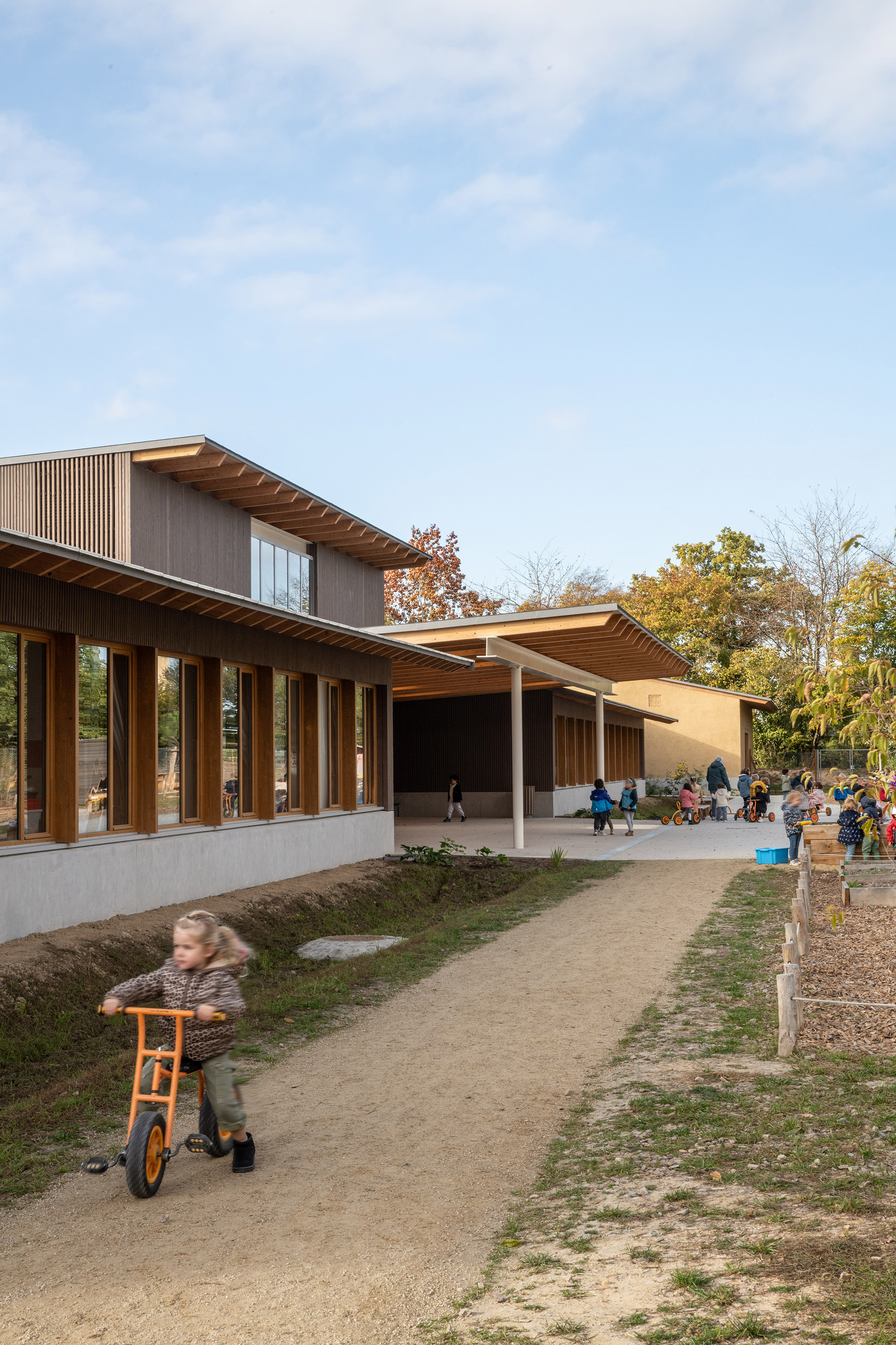 Exterior view with timber canopy over a concrete terrace where children play among autumn trees