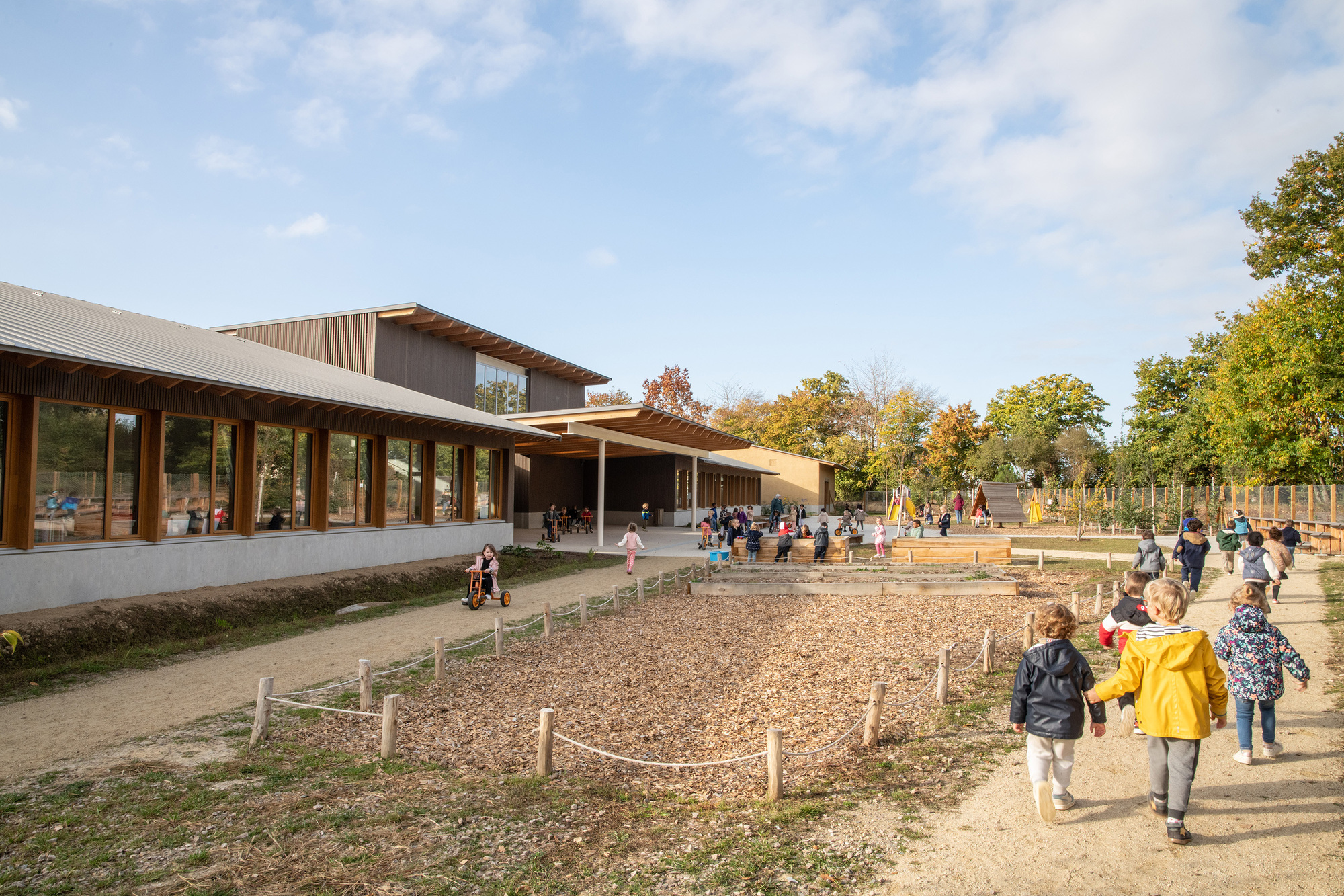 Courtyard entrance with children walking along gravel paths toward timber-clad volumes and playground beyond