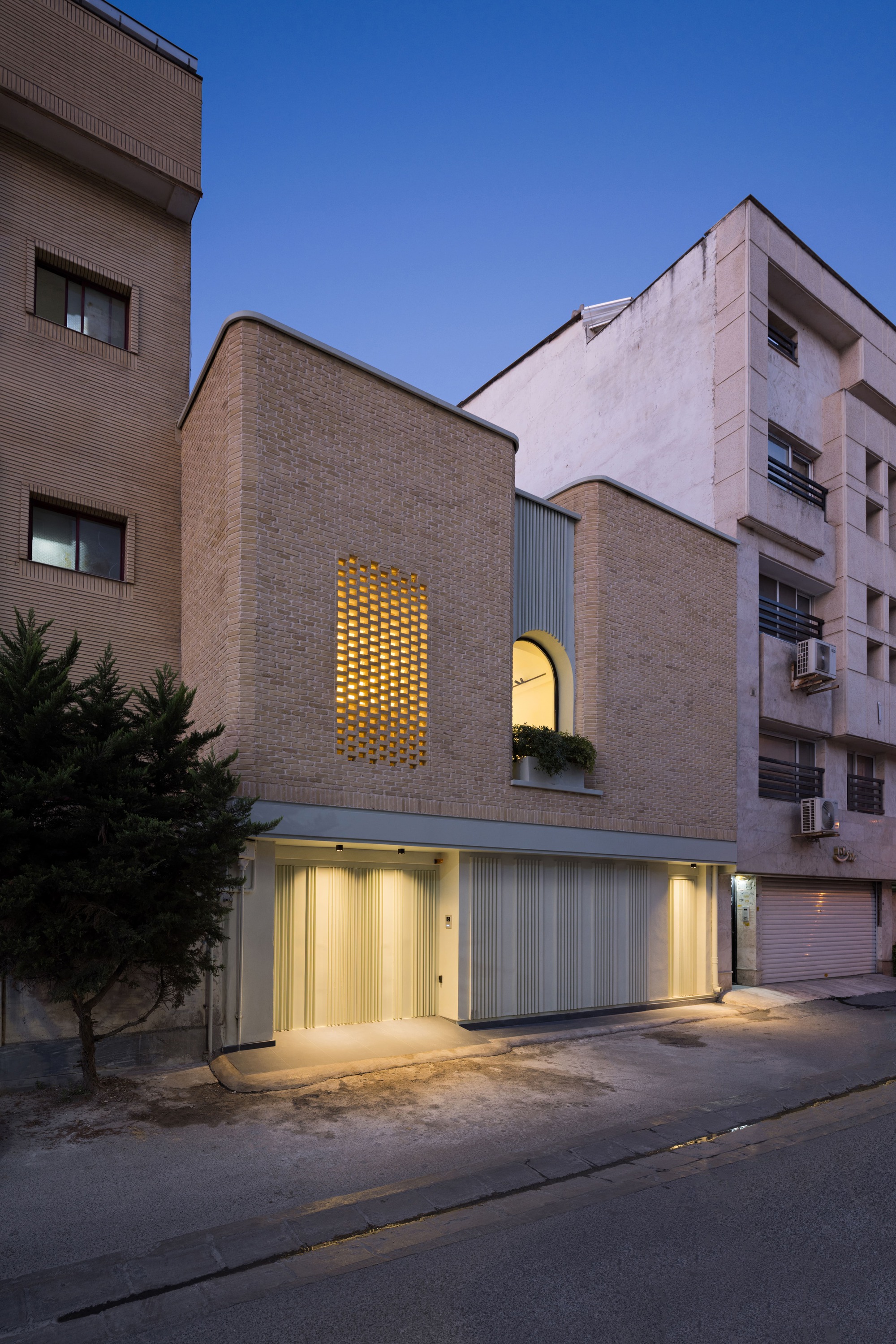 Brick facade with arched upper window and perforated panel illuminated at dusk between neighboring buildings