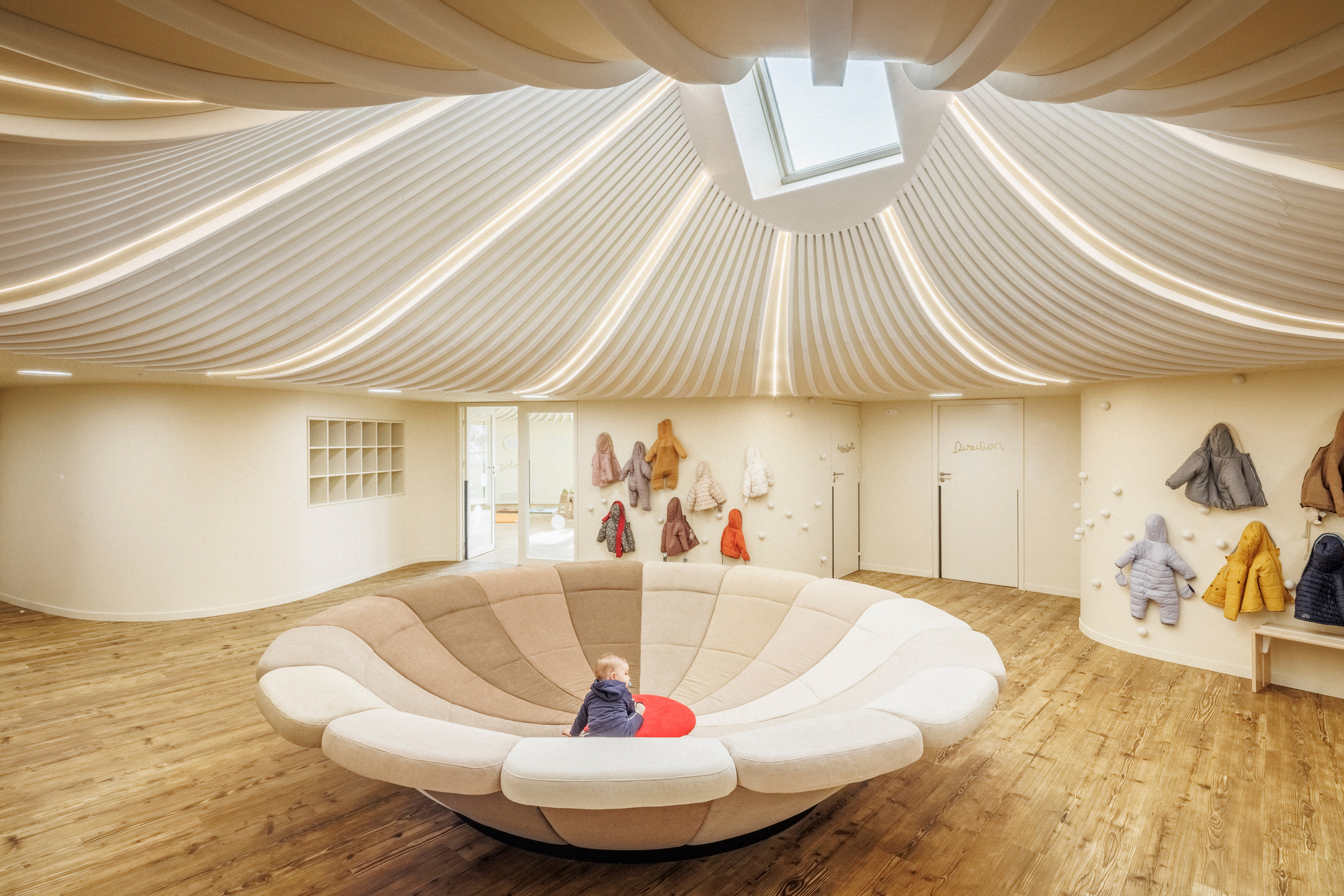 Interior playroom with upholstered circular seating platform beneath a sculptural timber ceiling and central skylight
