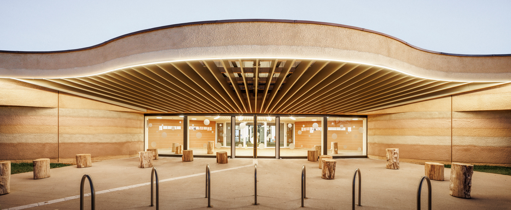 Evening view of the illuminated entrance with exposed timber rafters beneath the curved rammed earth canopy
