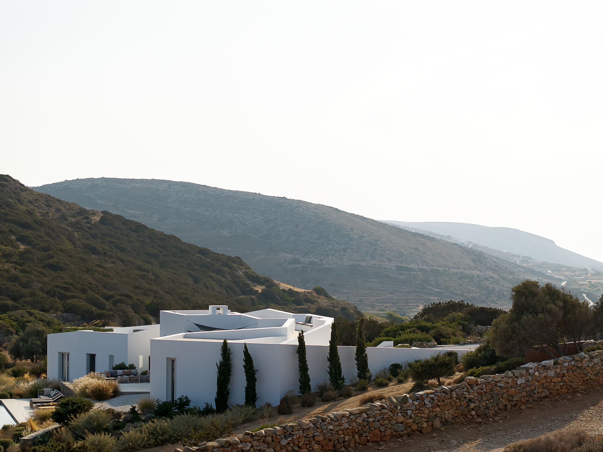 Side view of the white volumes with cypress trees against the mountain backdrop at dusk