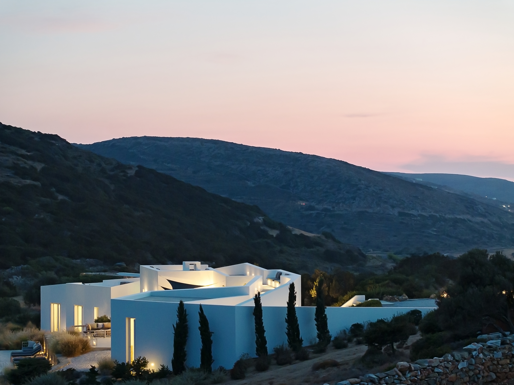 View of the clustered white volumes with uplighting and cypress trees at twilight