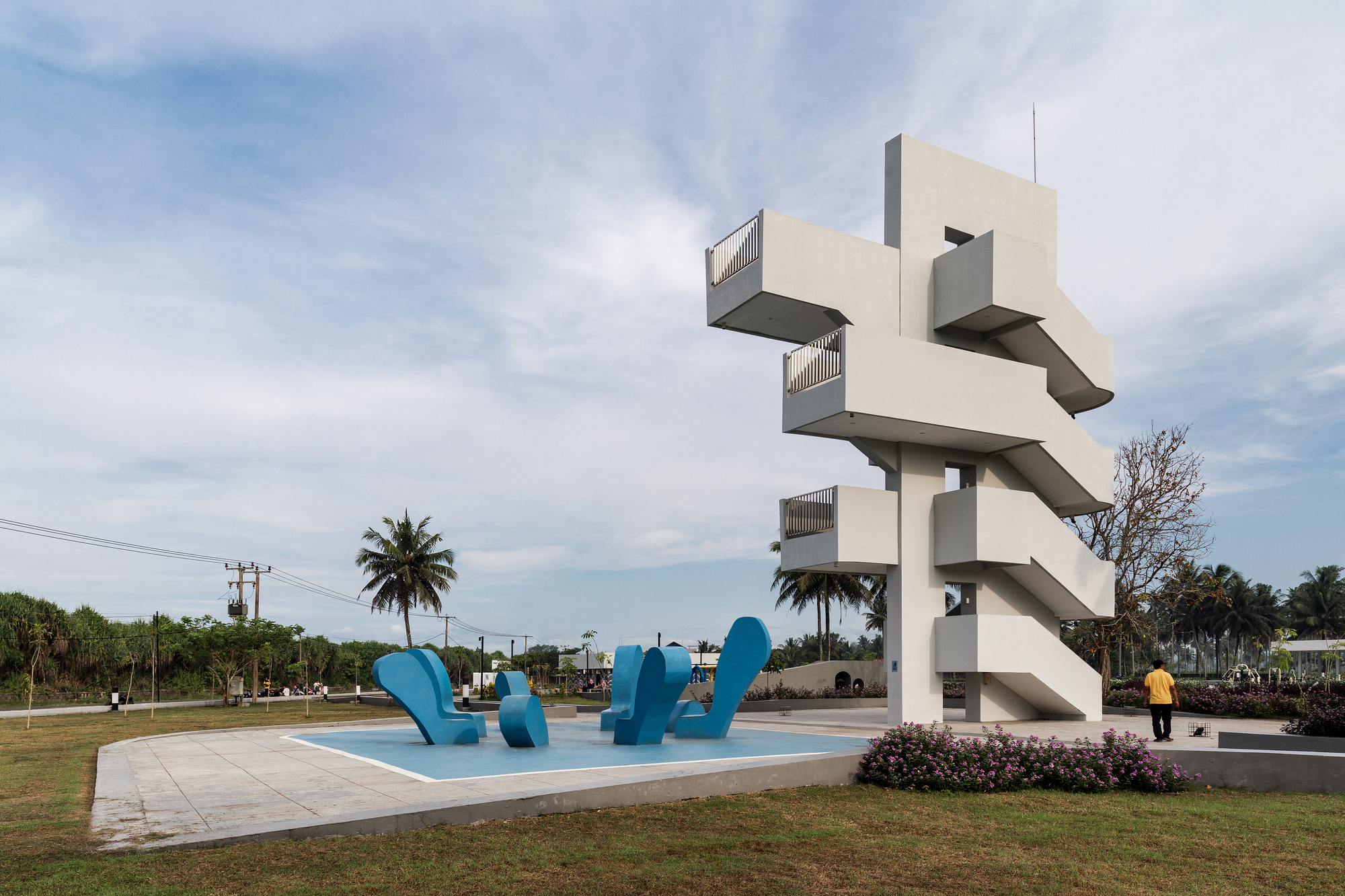 White concrete observation tower with cantilevered platforms beside blue sculptural figures on a plaza