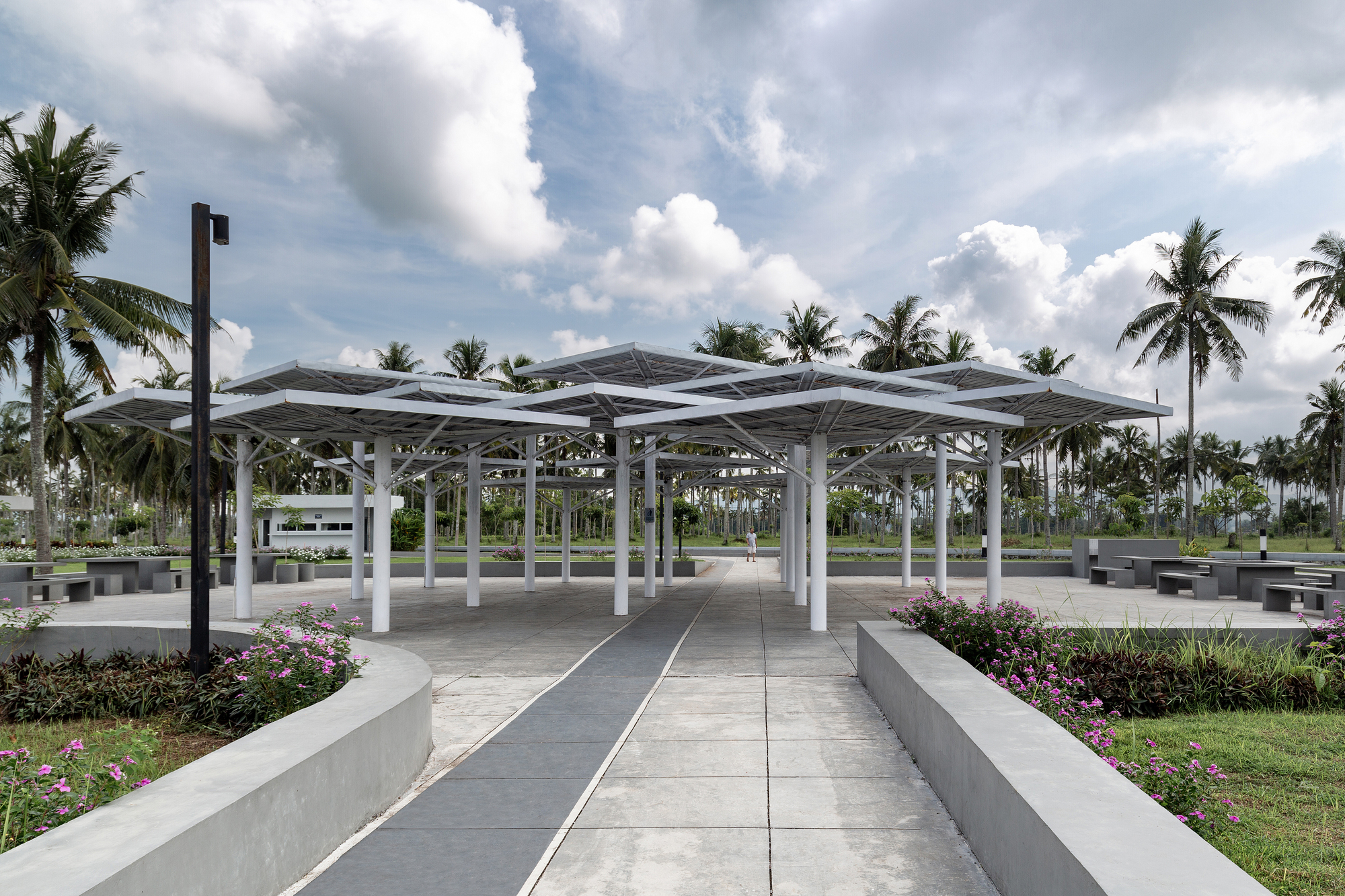 Open pavilion with folded white steel roof and circular columns framed by coconut palms
