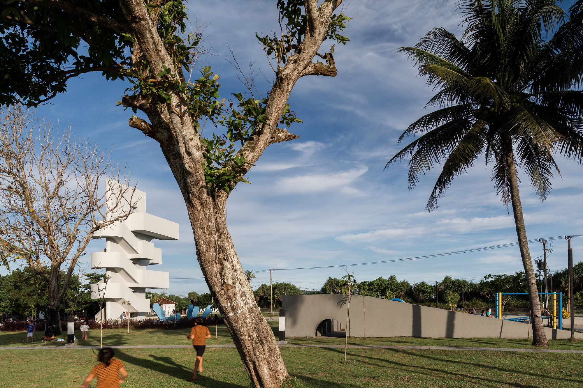 Park visitors walking past the white tower and curved concrete wall beneath palm trees