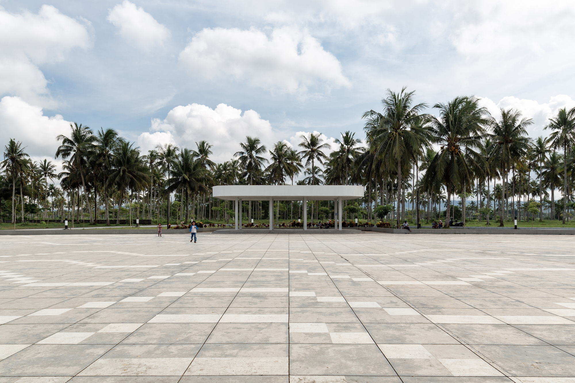 Wide paved plaza with large-format stone tiles leading to the slender-columned pavilion under a cloudy sky