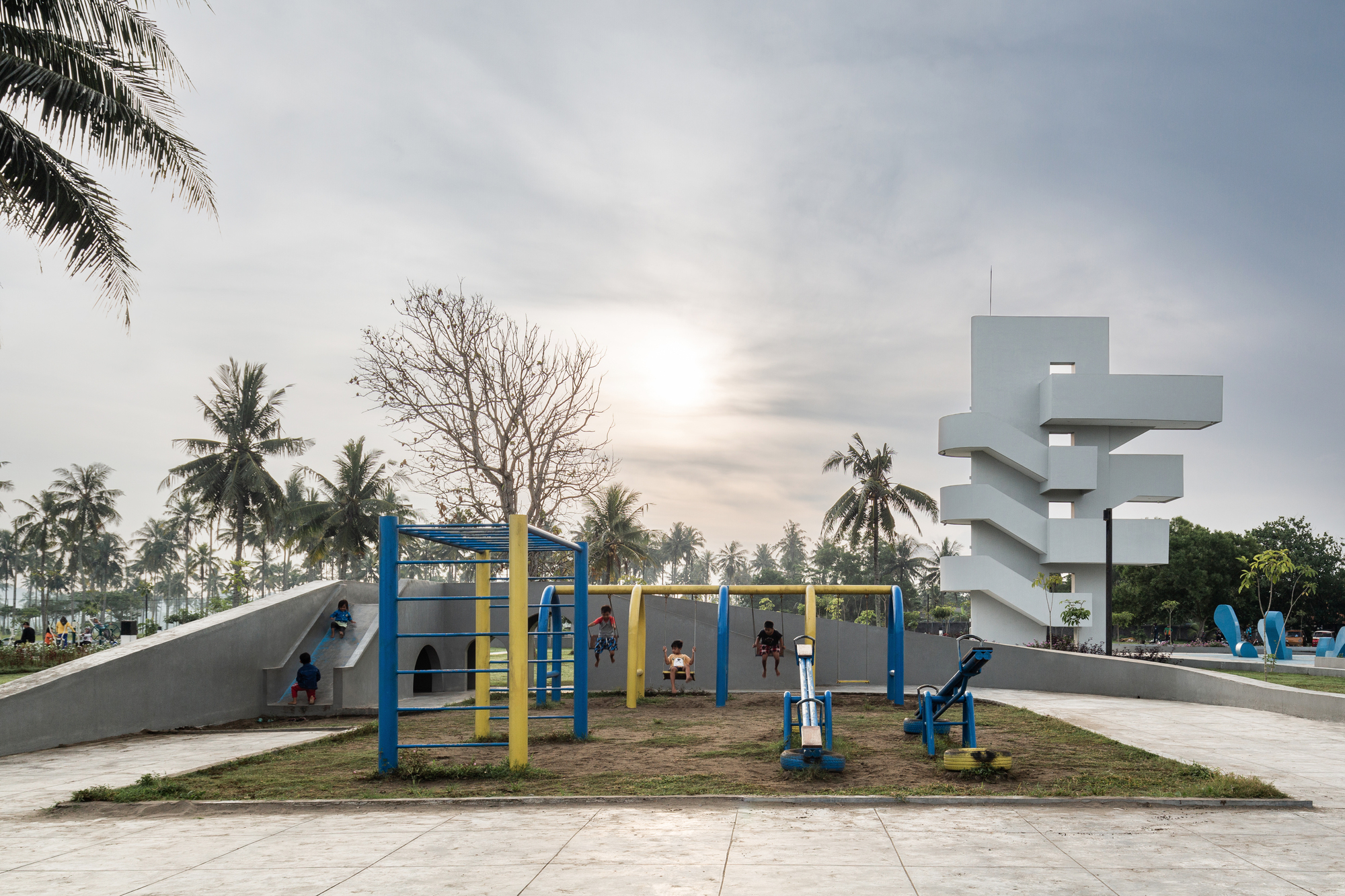 Children playing on blue and yellow playground equipment beside the white concrete tower at dusk