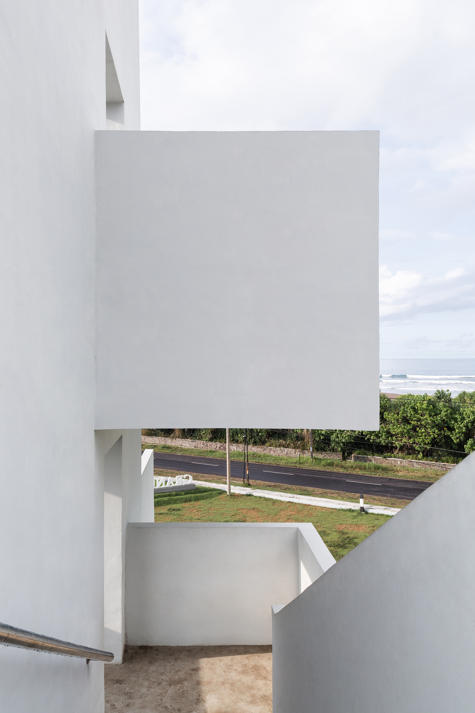View through white-walled stairwell opening showing distant ocean and roadway beyond palm trees
