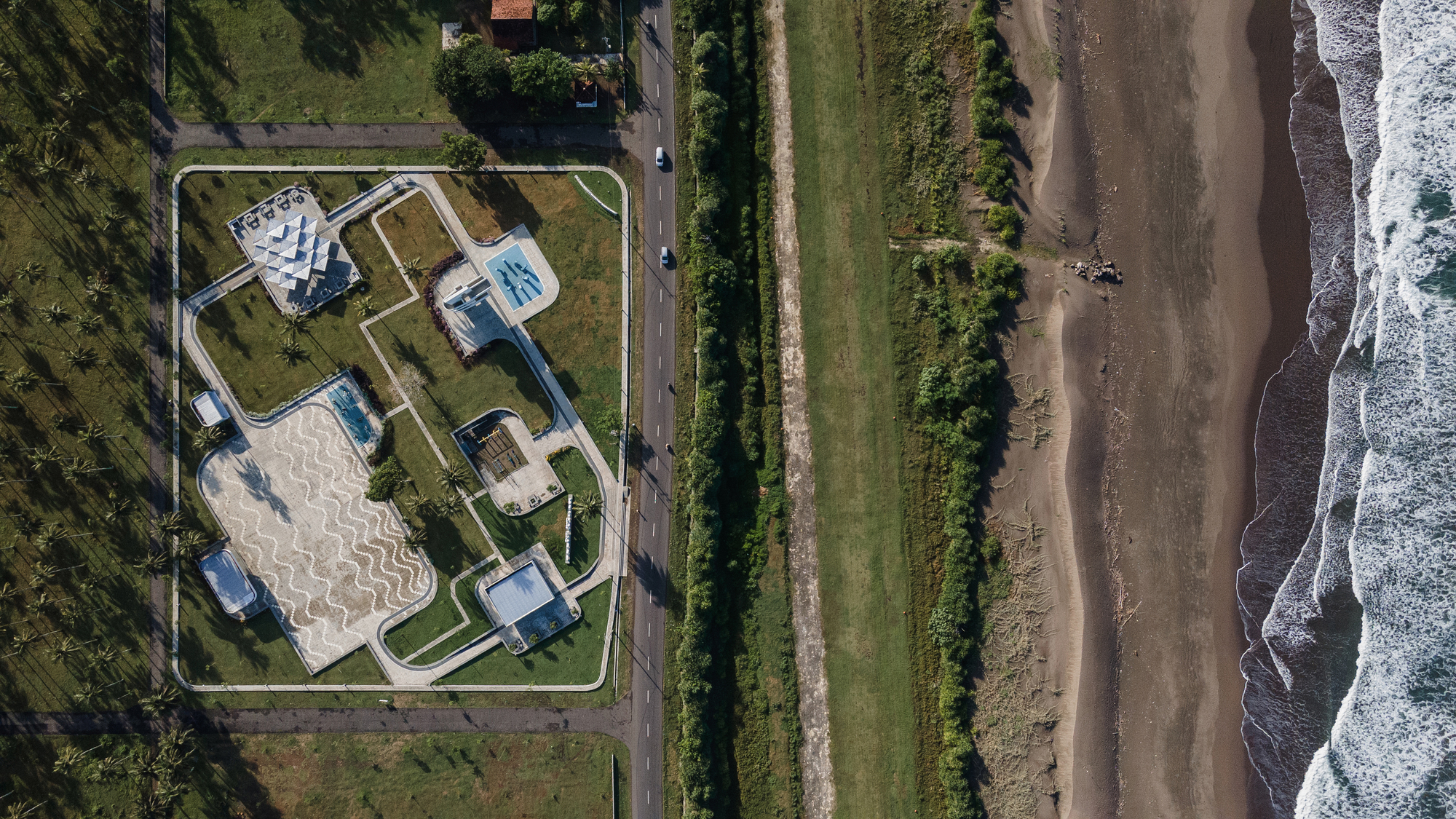 Aerial view of compound with white volumes and serpentine paving adjacent to coastal road and beach