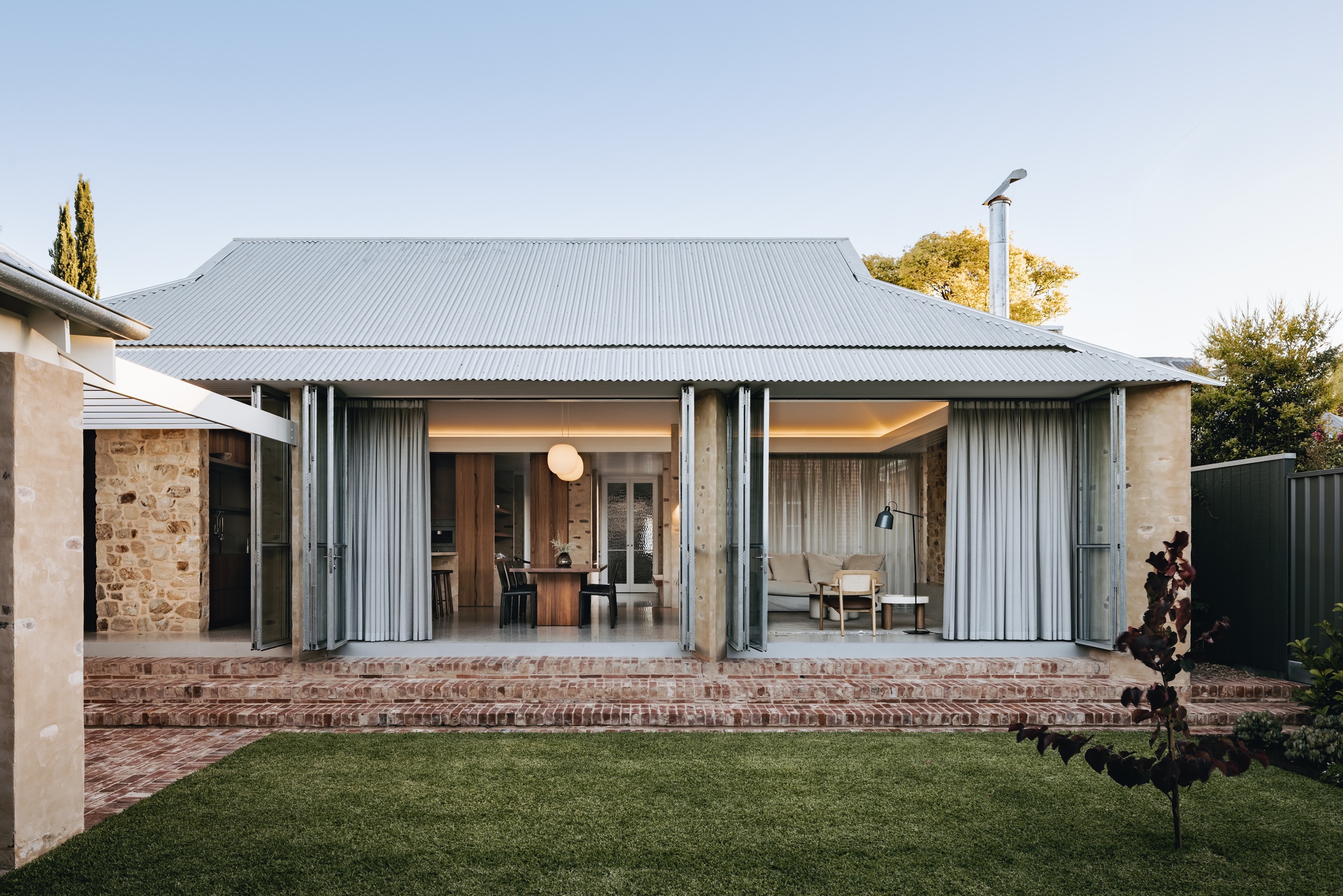 Rear elevation with corrugated metal roof and folding glass doors opening onto brick steps and lawn