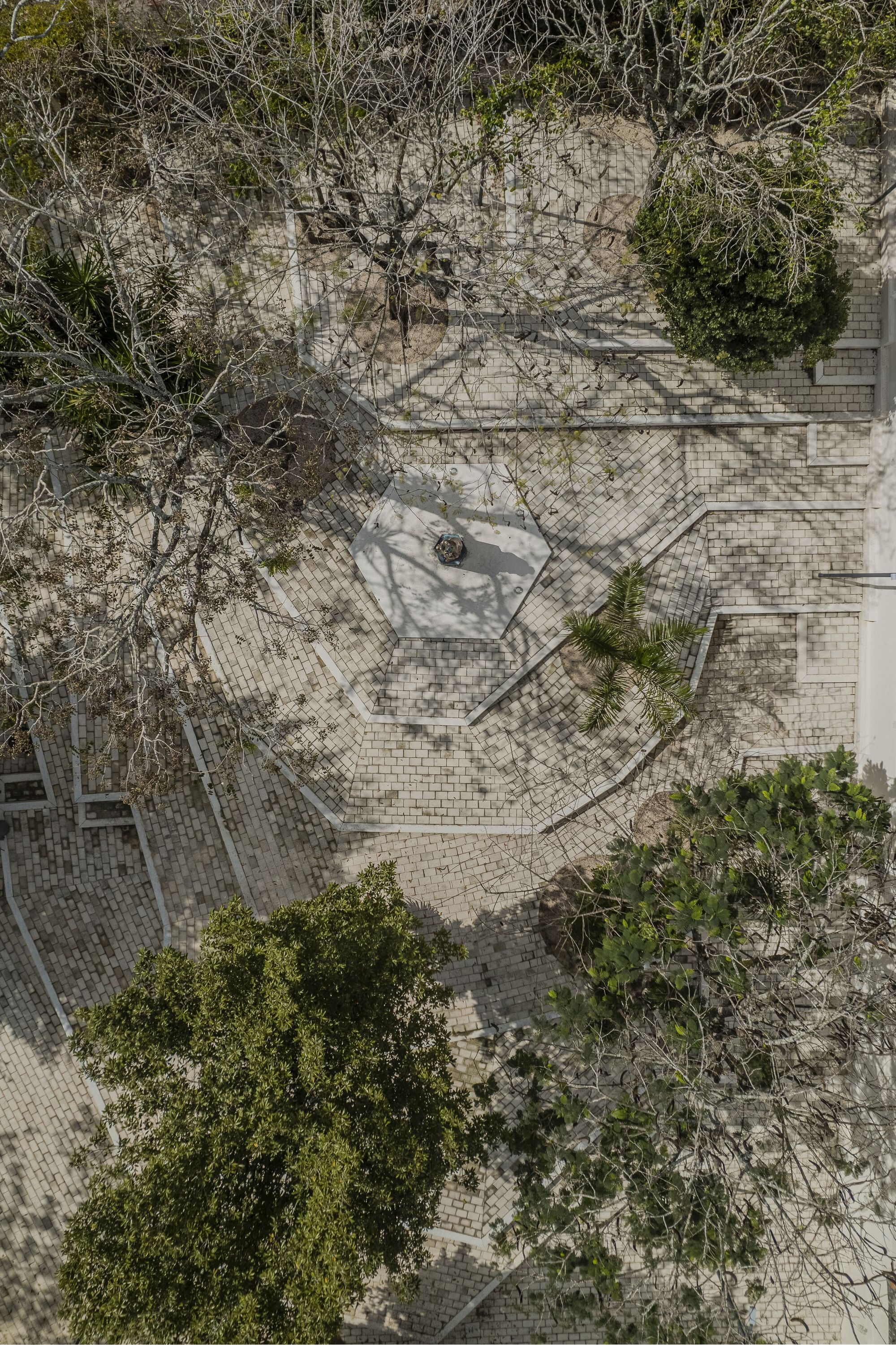 Drone view of stone-paved plaza with diagonal pathways, scattered trees, and a single person walking