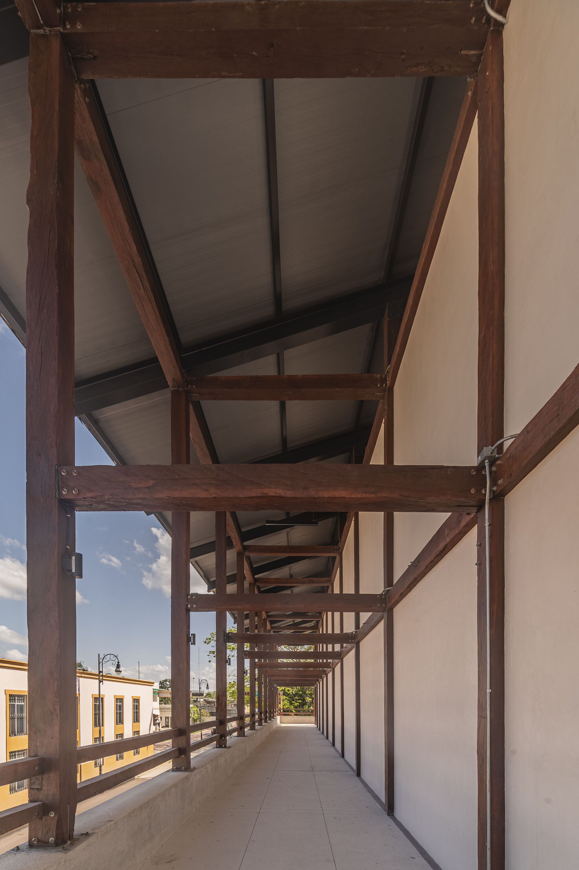 Exterior elevated walkway with timber frame structure and translucent roof panels overlooking neighboring buildings